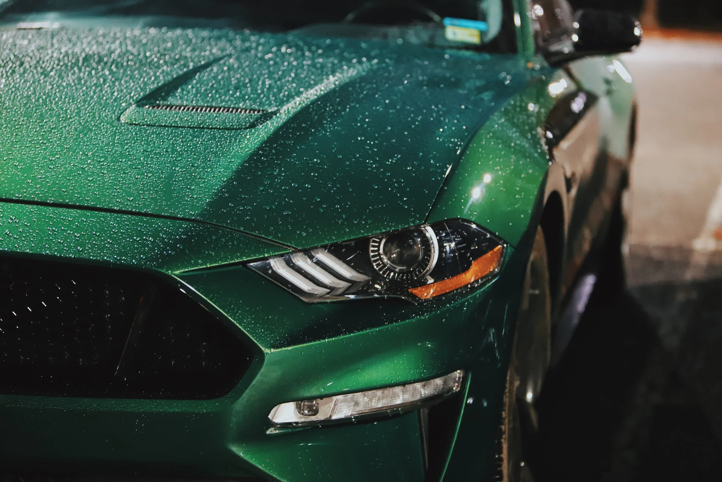Close-up of a green sports car with rain droplets on its surface, featuring sleek headlight design and aerodynamic curves.