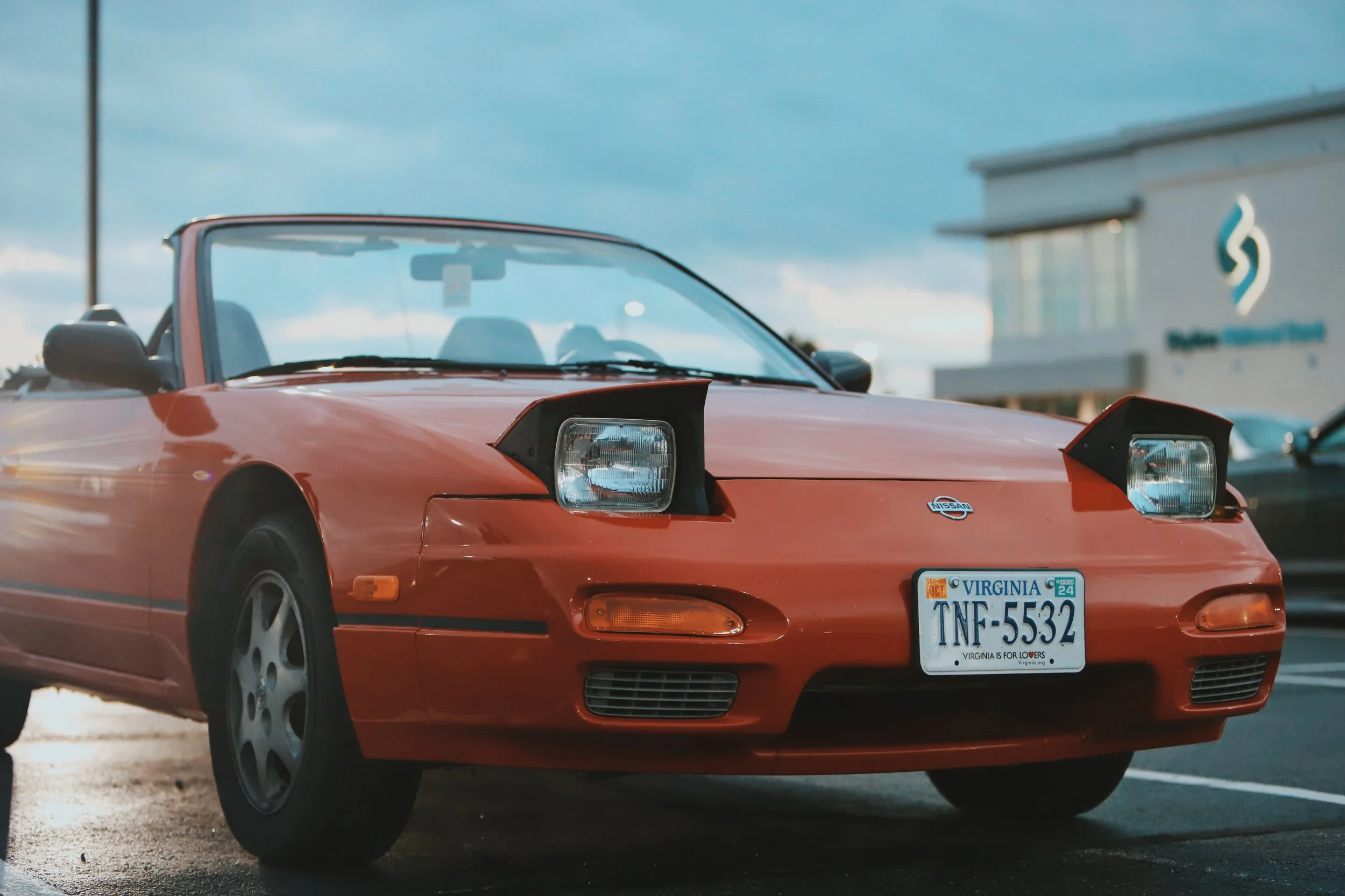 Red Nissan convertible car with raised pop-up headlights, parked in a lot near a building, under a cloudy sky.