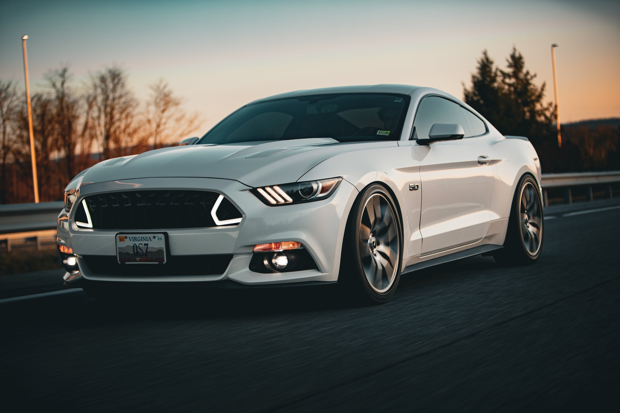 A white Ford Mustang sports car driving on a highway at sunset with trees and sunset sky in background.