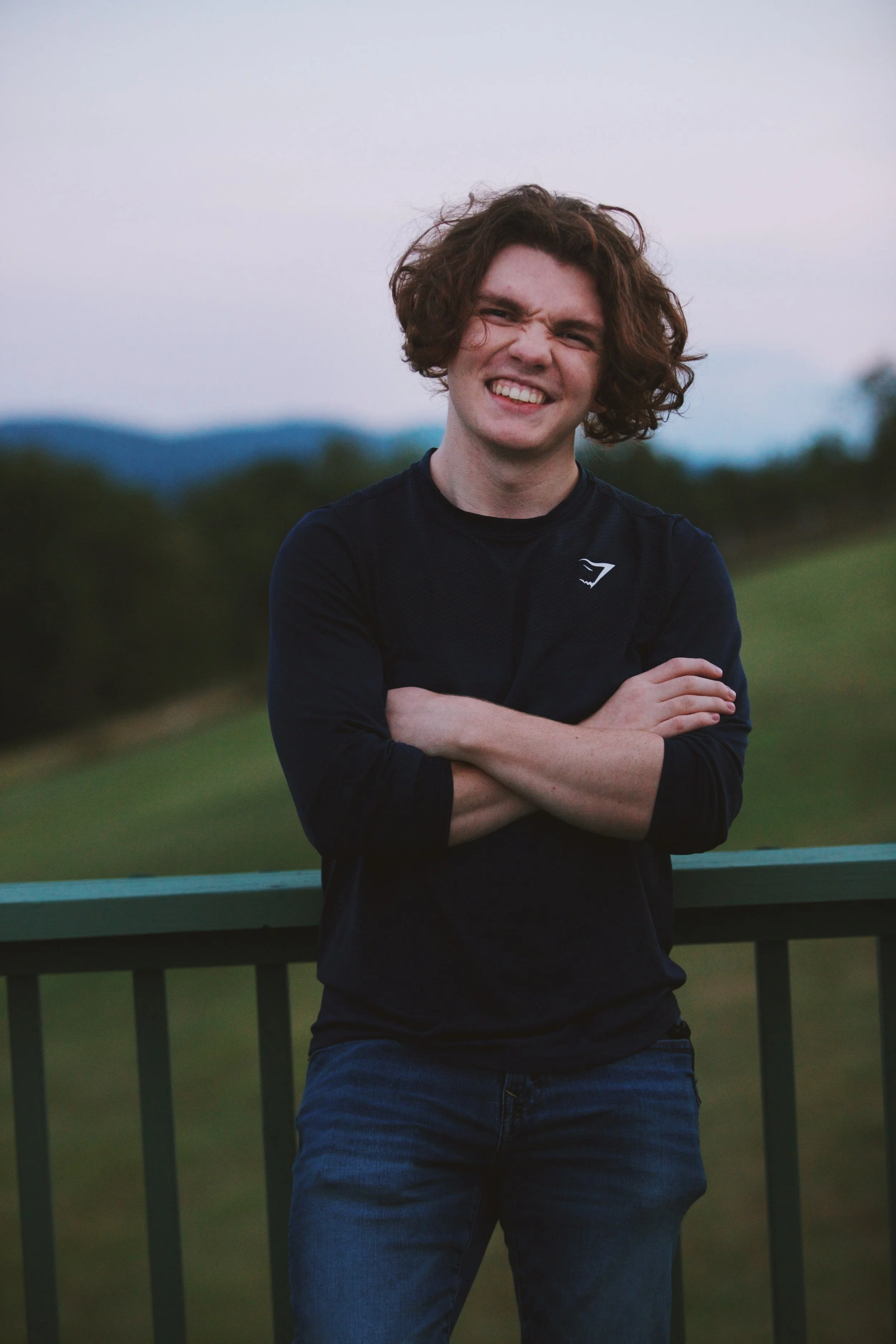 A young man with curly brown hair smiling and crossing his arms, standing outdoors on a railing with a blurred landscape of trees and mountains in the background.