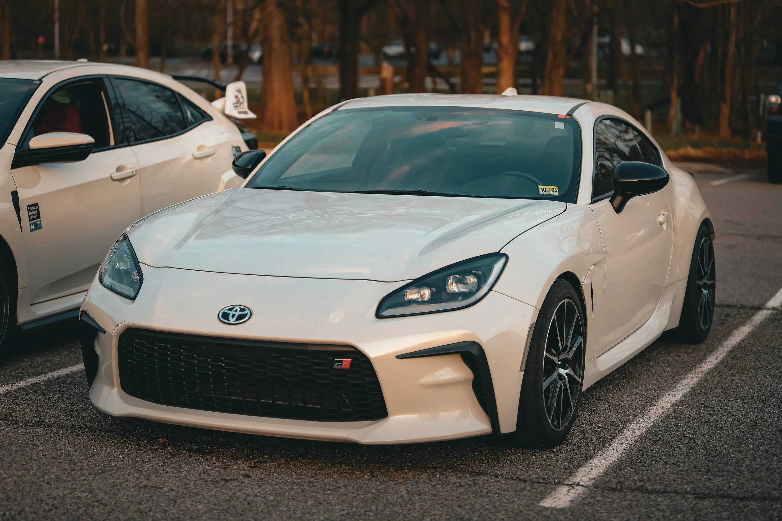 A white Toyota sports car parked in a parking lot next to another white car, with trees in the background, during golden hour.