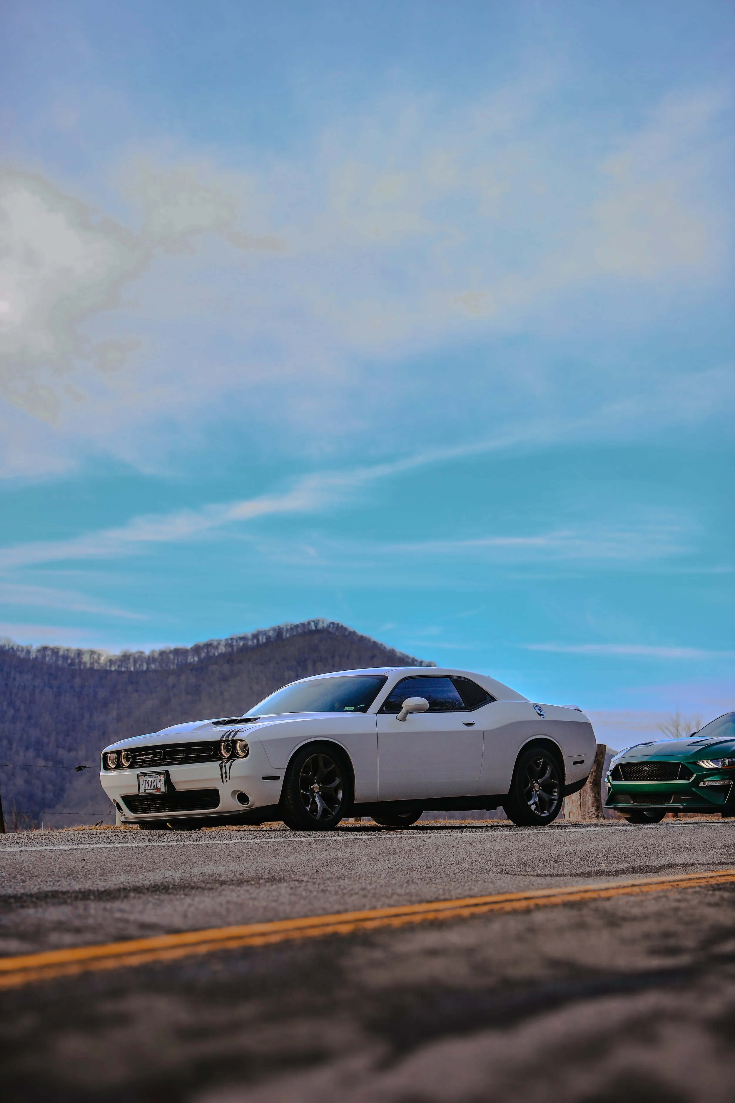 A white sports car parked on the side of a road with mountains in the background and a partly cloudy sky overhead.