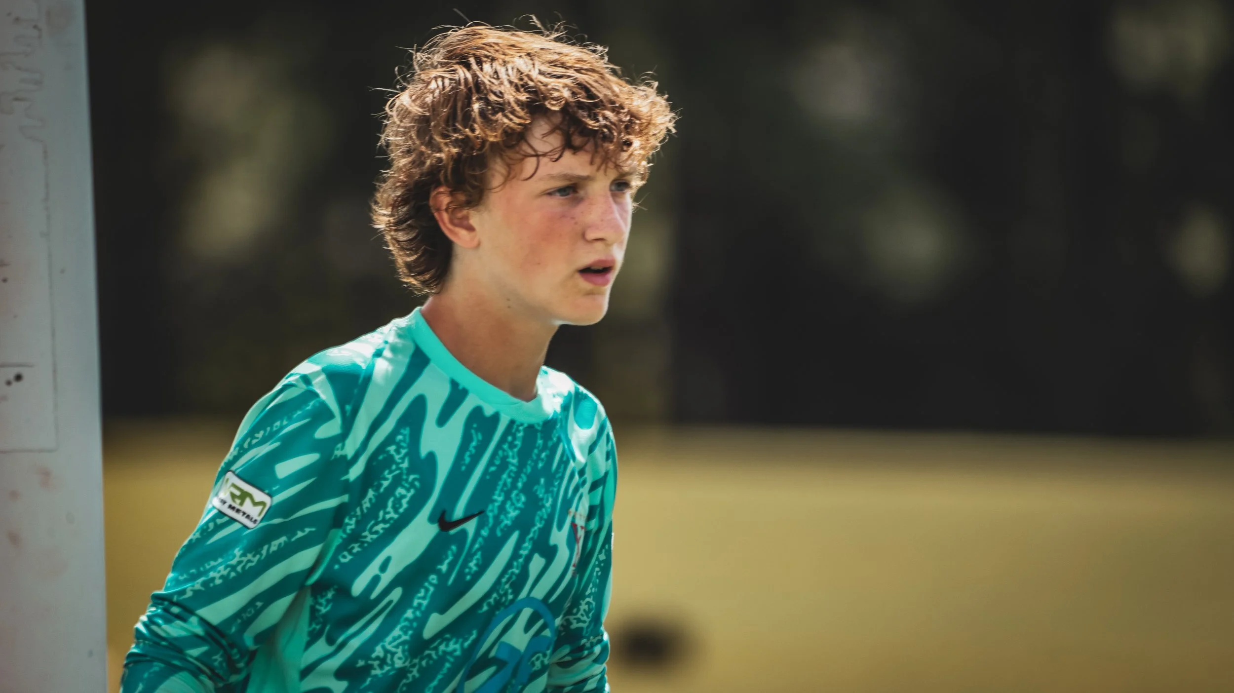 Young male soccer goalkeeper standing on the field, wearing a teal patterned long-sleeve jersey, with a focused expression, outdoors with blurred background of trees and grass.