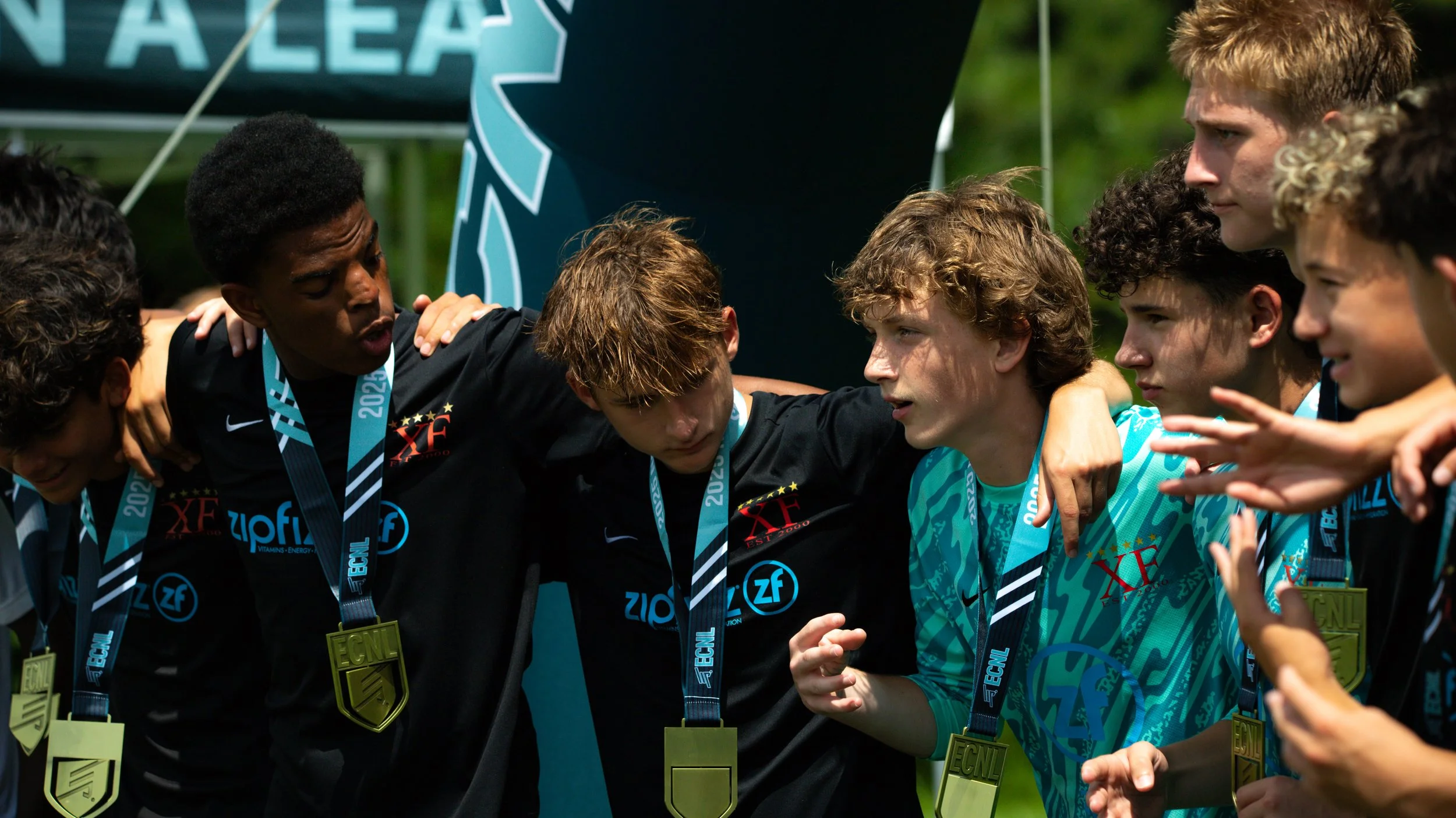 Young soccer players huddled together after a game, wearing medals and team jerseys, with their arms around each other's shoulders.