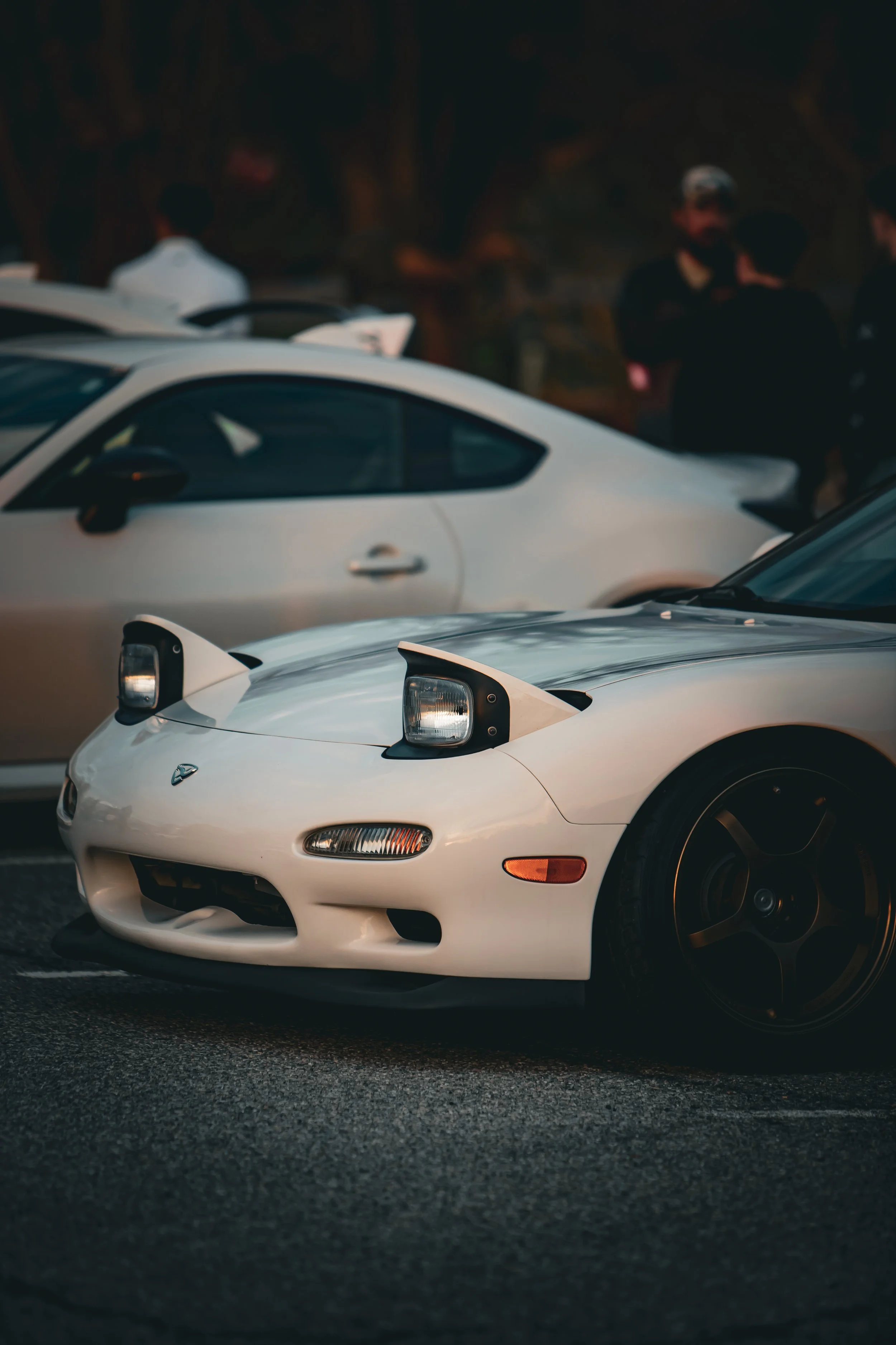 A white Mazda RX-7 sports car with pop-up headlights in the foreground and a silver coupe in the background, with people standing and talking behind the cars.