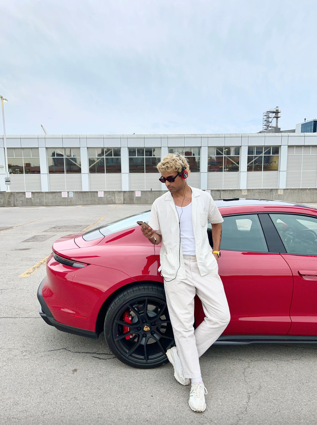 Art directed automotive lifestyle campaign featuring a male talent in an all-white outfit leaning against a red Porsche Taycan in an urban parking lot setting.