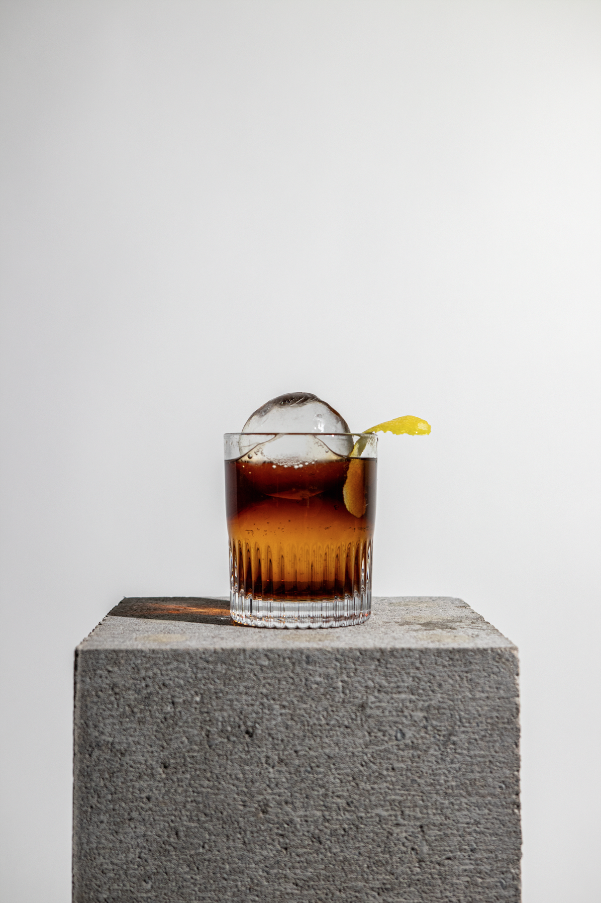 Art directed product photography of a dark cocktail with large ice sphere and citrus garnish on a concrete pedestal, created for Ice-ASAP.