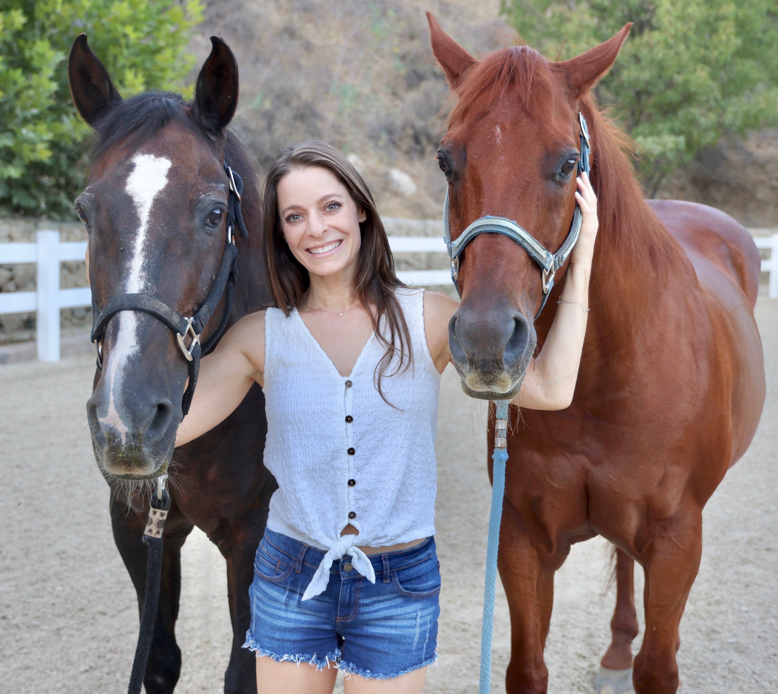 A woman with brown hair and a white top smiling while holding the heads of two horses, one black and one brown, in an outdoor setting with a white fence and green trees in the background.