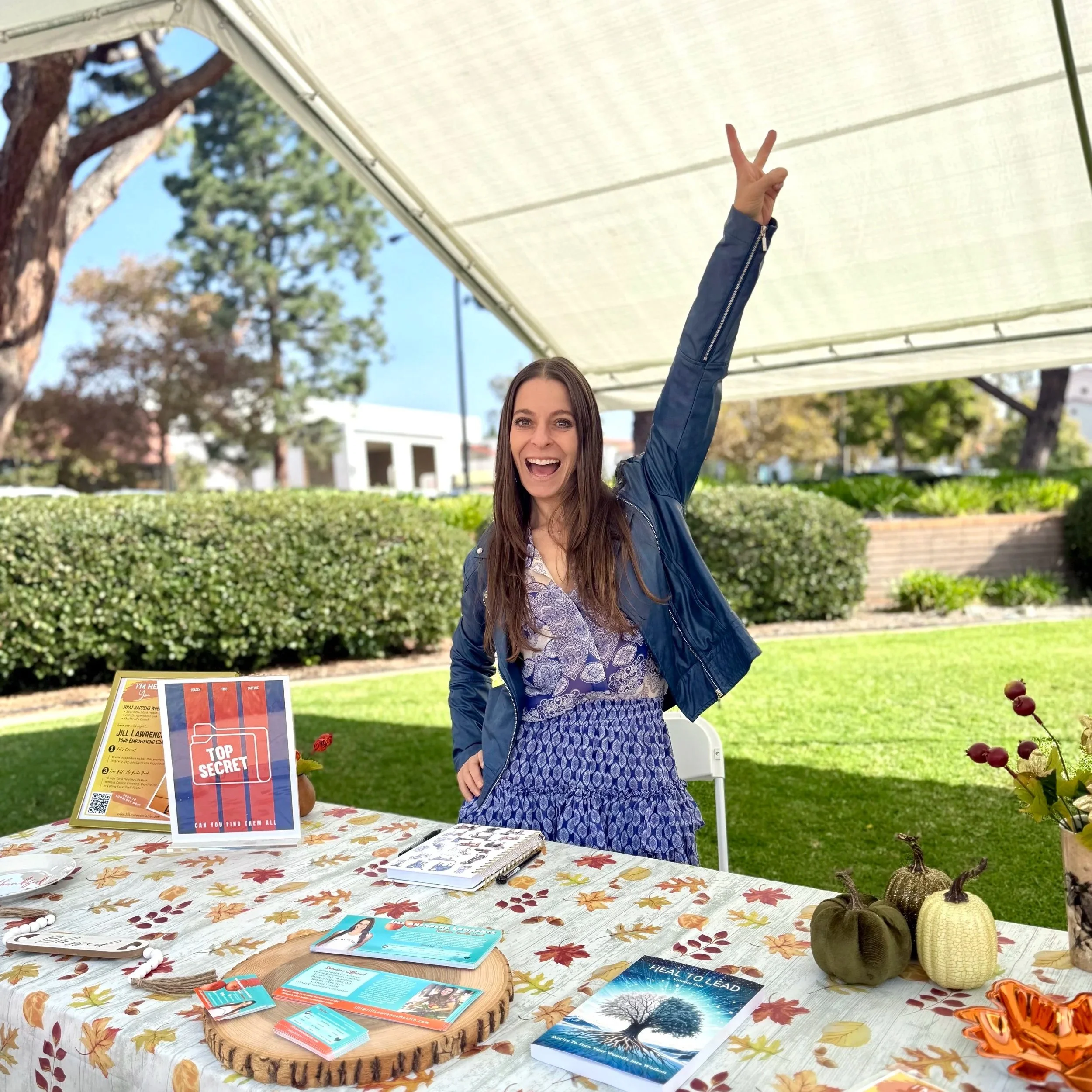 A smiling woman with long brown hair wearing a blue jacket and patterned dress, holding her hand up making a peace sign, standing behind a table with informational brochures, pamphlets, and fall-themed decorations outdoors under a canopy.