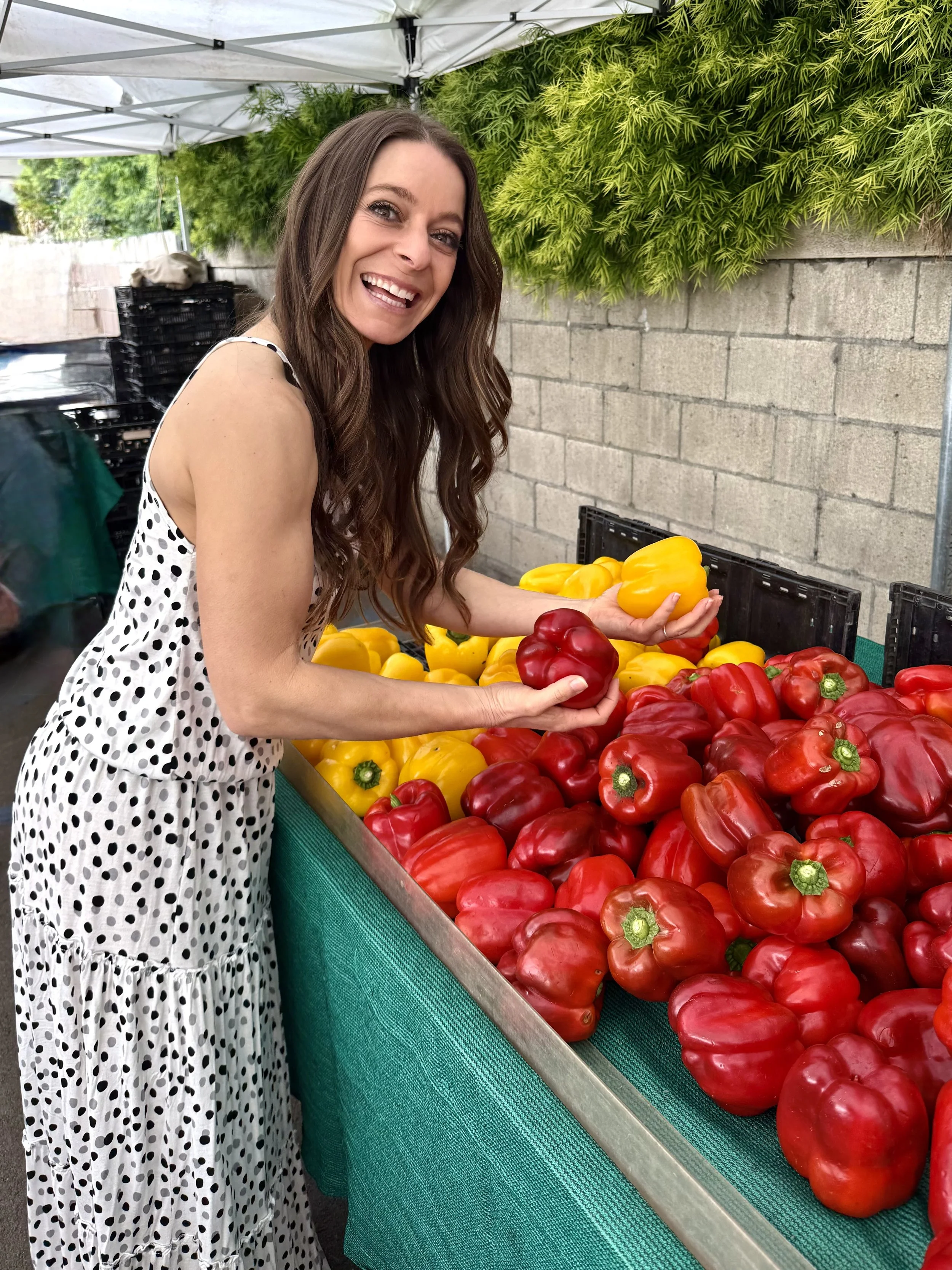 A woman with long brown hair wearing a white dress with black polka dots is shopping for bell peppers at an outdoor market. She is smiling and holding a red bell pepper. The market stall has red and yellow bell peppers displayed on a green tablecloth, with a brick wall and greenery in the background.