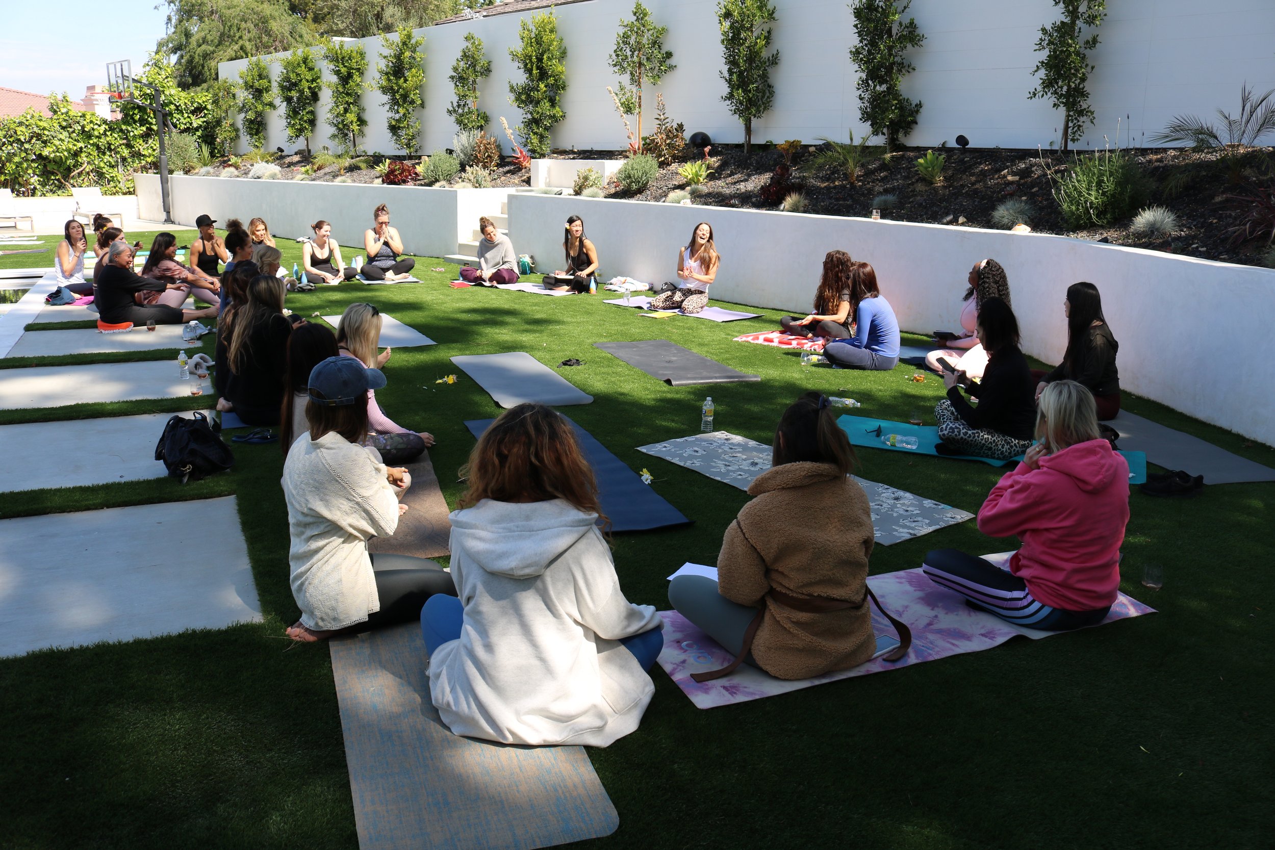 A group of women seated on yoga mats in a circle on a grassy outdoor area, participating in a yoga or meditation class on a sunny day.