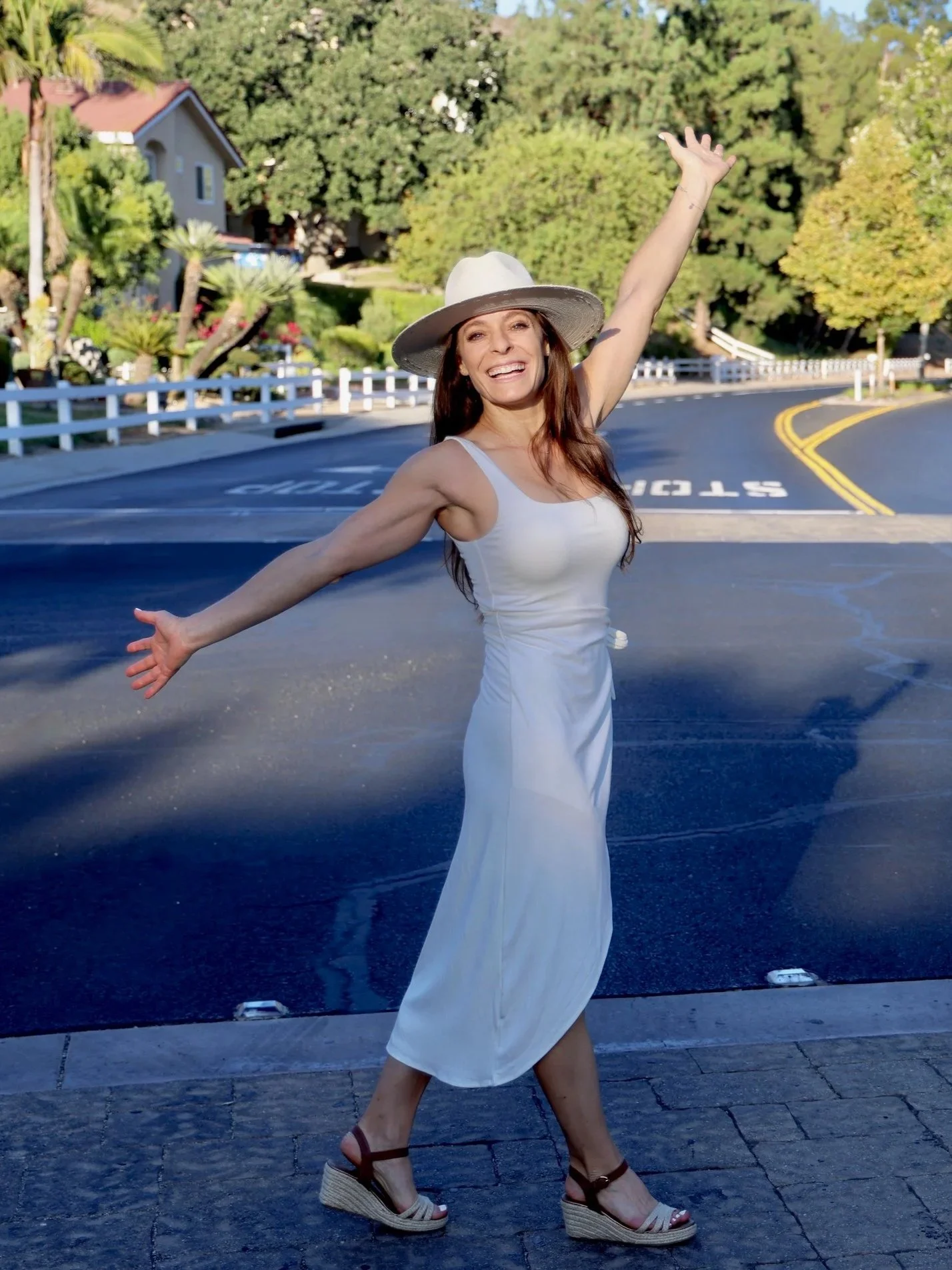 A woman wearing a white sleeveless dress and a wide-brimmed hat, smiling with arms outstretched, standing on a street with trees and houses in the background.