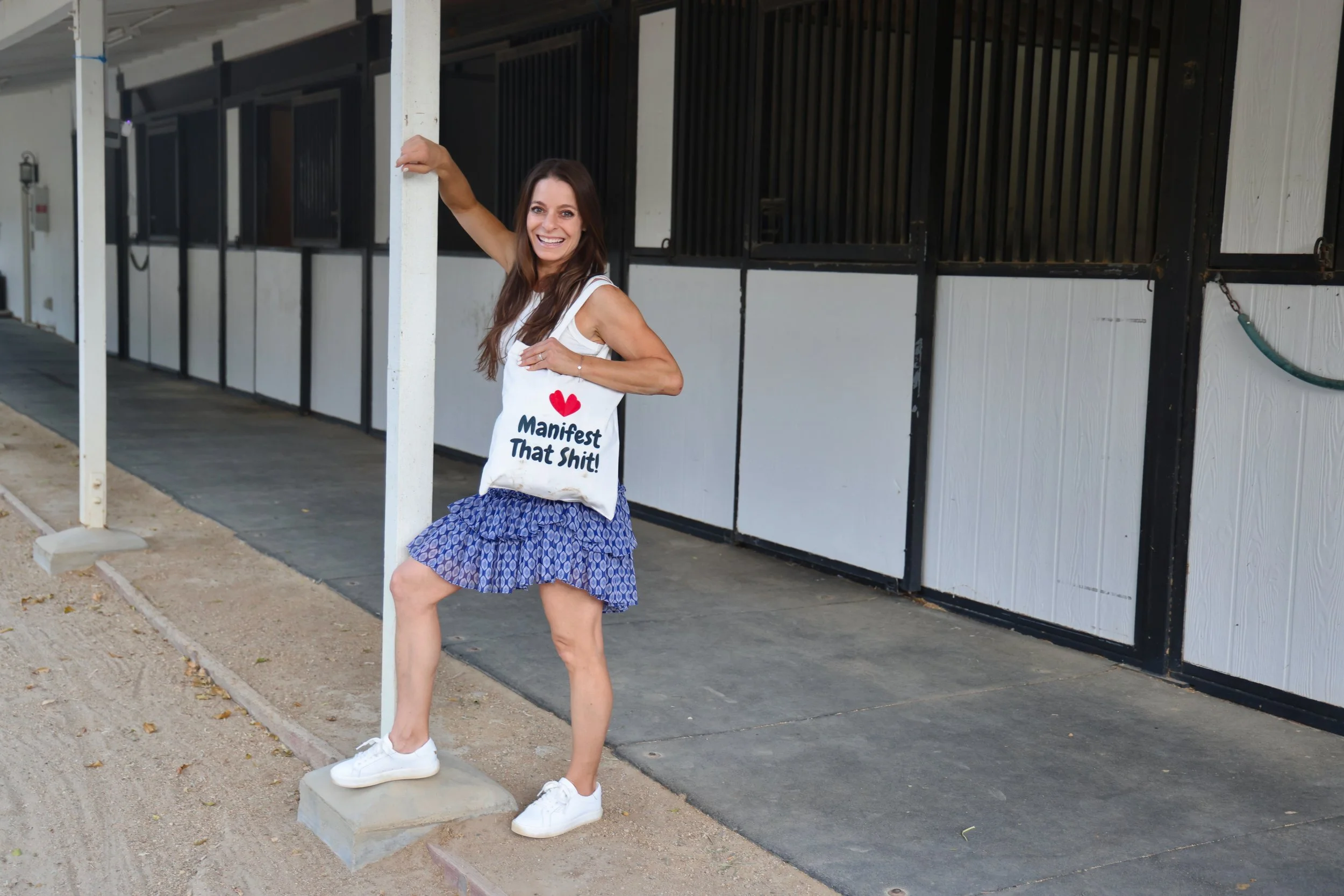 Woman smiling and posing with one foot on a concrete block, holding onto a vertical post, standing in front of a stable or barn with black and white walls.