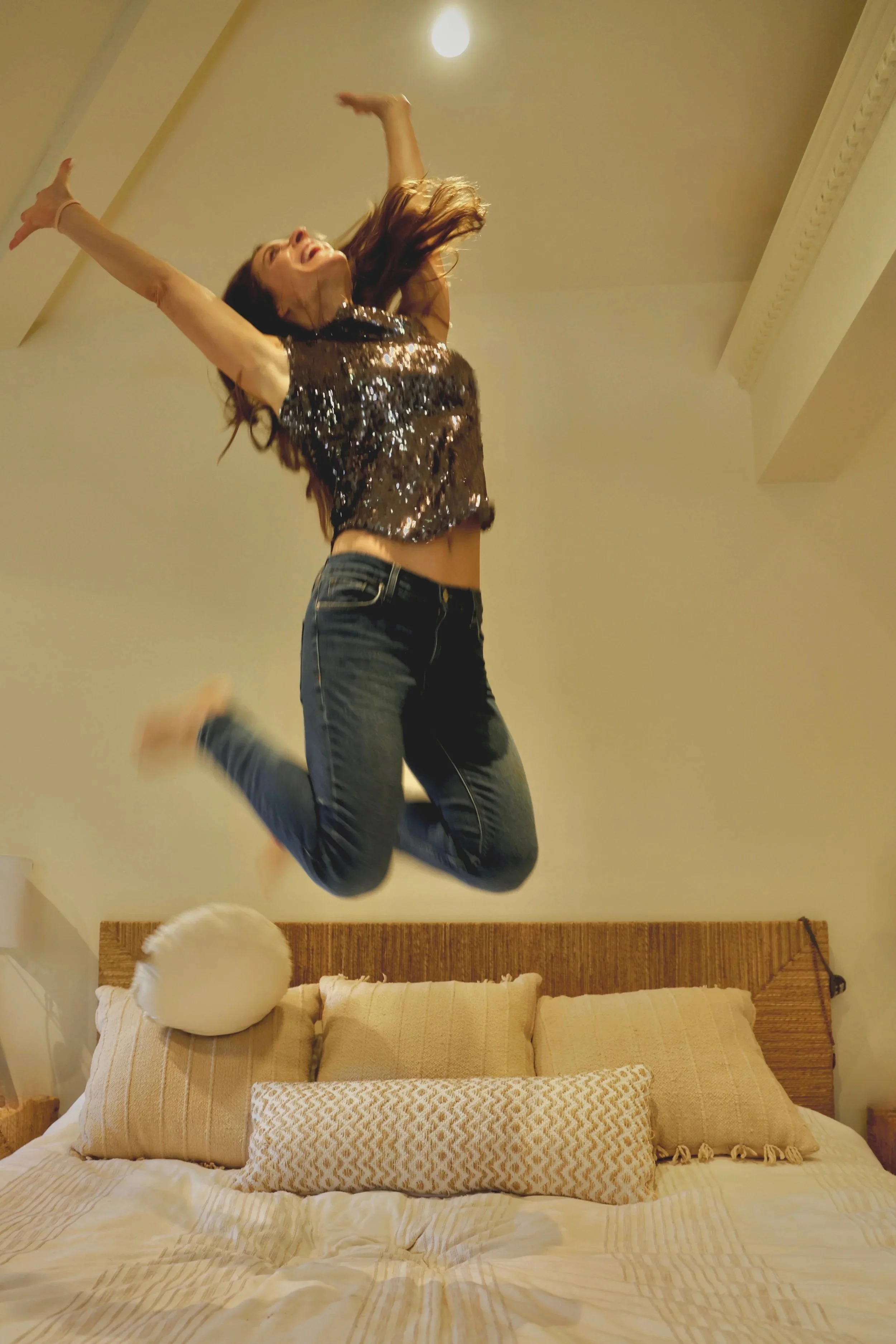 A woman is jumping on a bed in a bedroom. She is smiling, wearing a sparkly top and jeans, and has long hair flowing as she leaps.