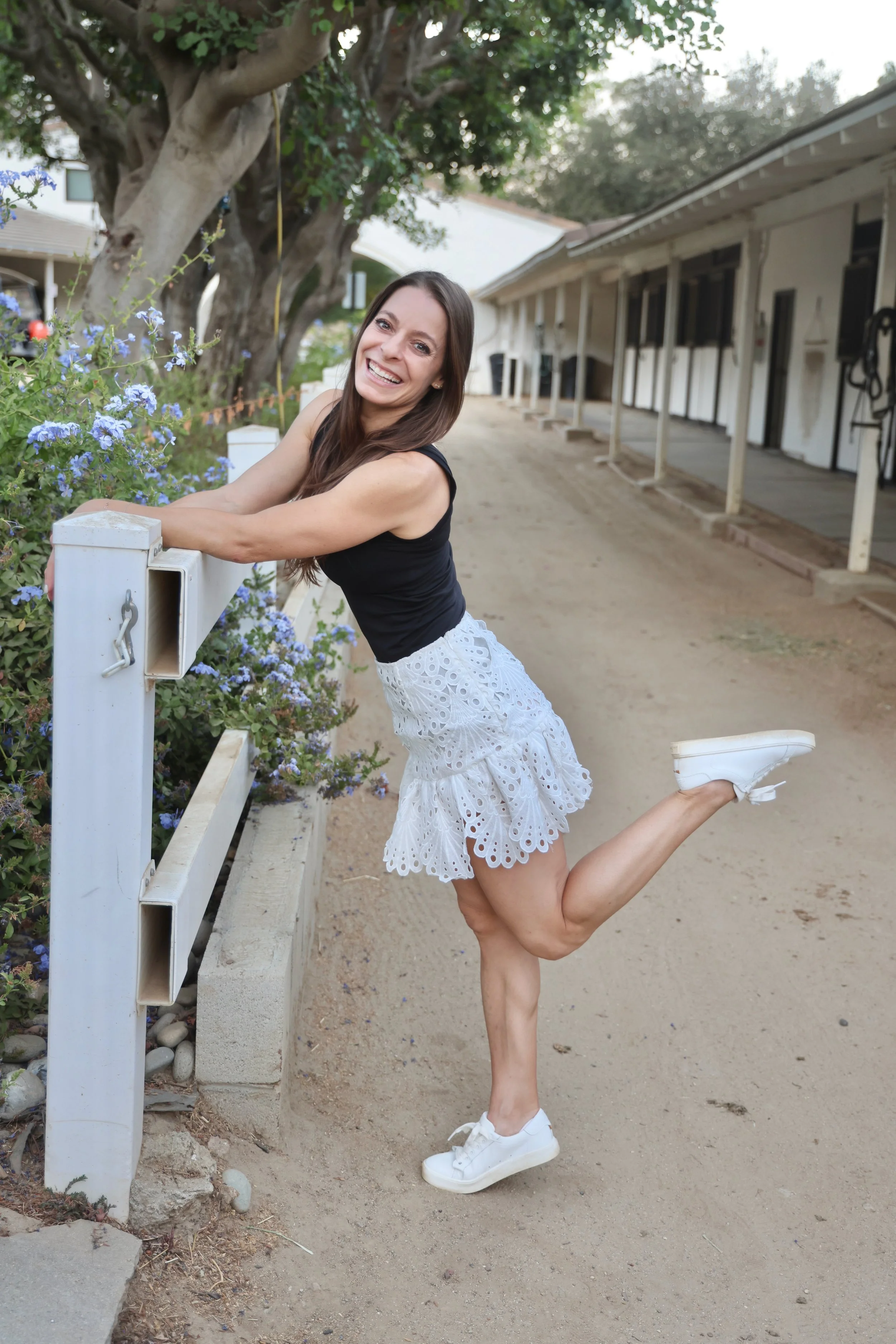 A woman in a black top and white eyelet skirt kneeling on one leg outdoors, smiling and holding onto a white fence with purple flowers nearby.