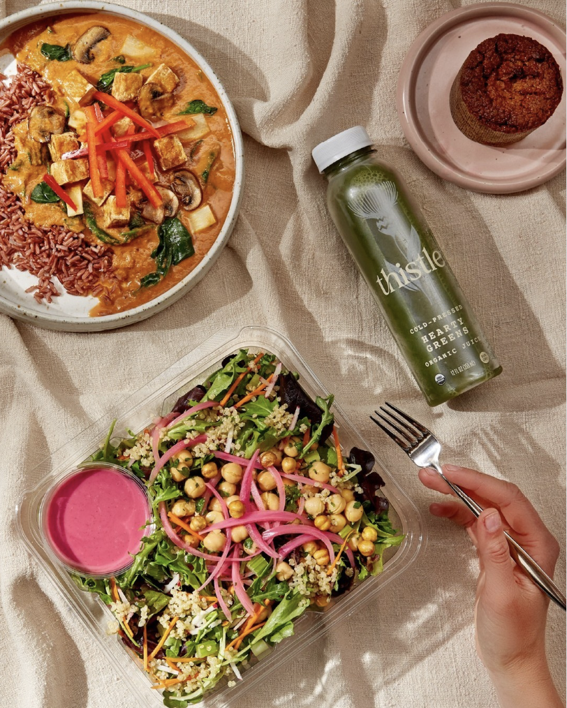 A top-down view of a meal setting on a beige cloth, including a bowl of vegetable curry with brown rice, a plastic container of mixed greens salad with chickpeas, pickled onions, and a pink dressing, a bottle of green cold-pressed juice labeled 'thistle' with leafy design, a pink muffin on a small round pink plate, and a hand holding a fork.