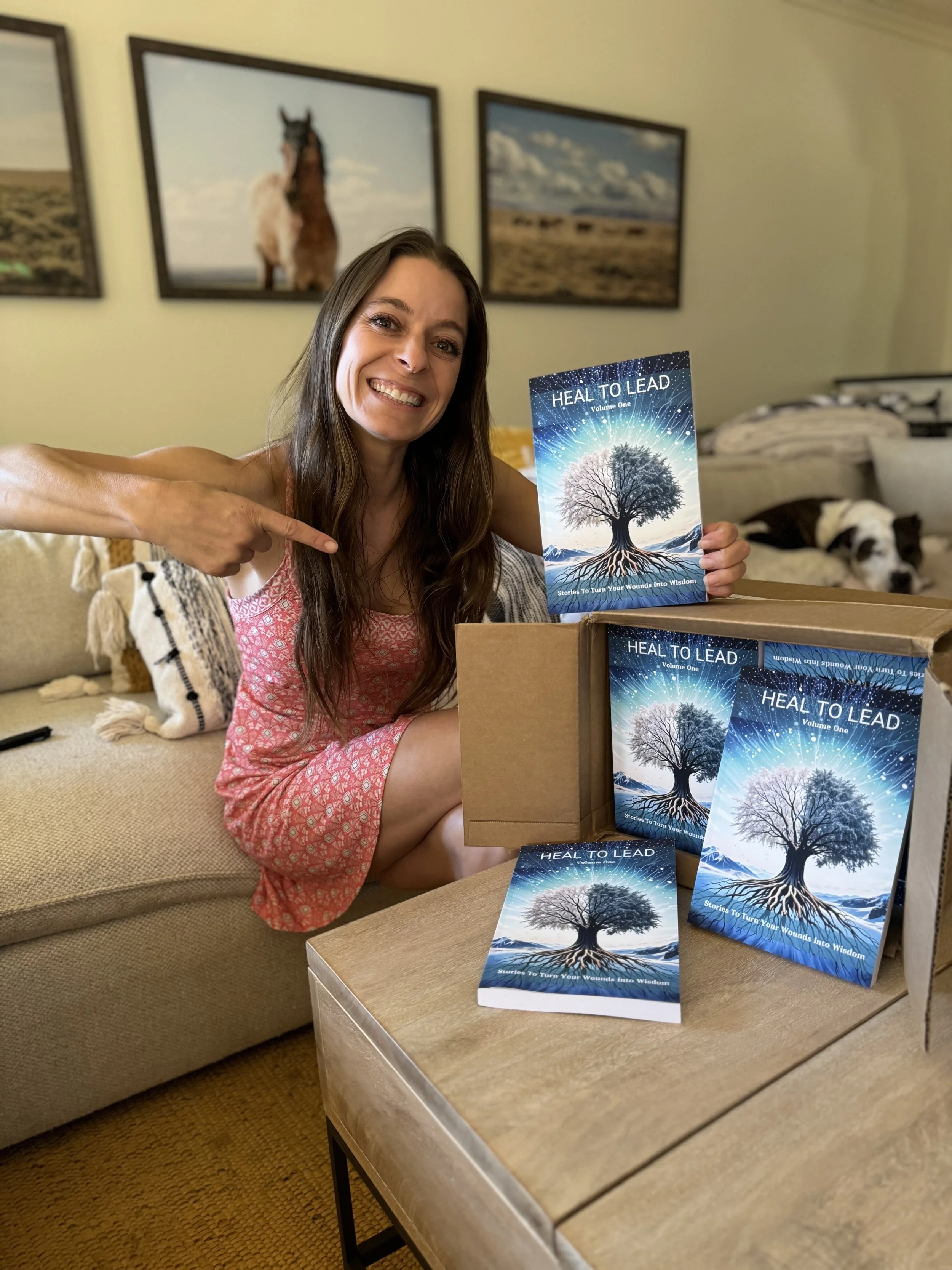 Woman sitting on a beige couch holding a book titled 'Heal to Lead' with a tree with roots illustration, while pointing at it, with dog resting on the couch behind her, and framed pictures of horses and landscapes on the wall.