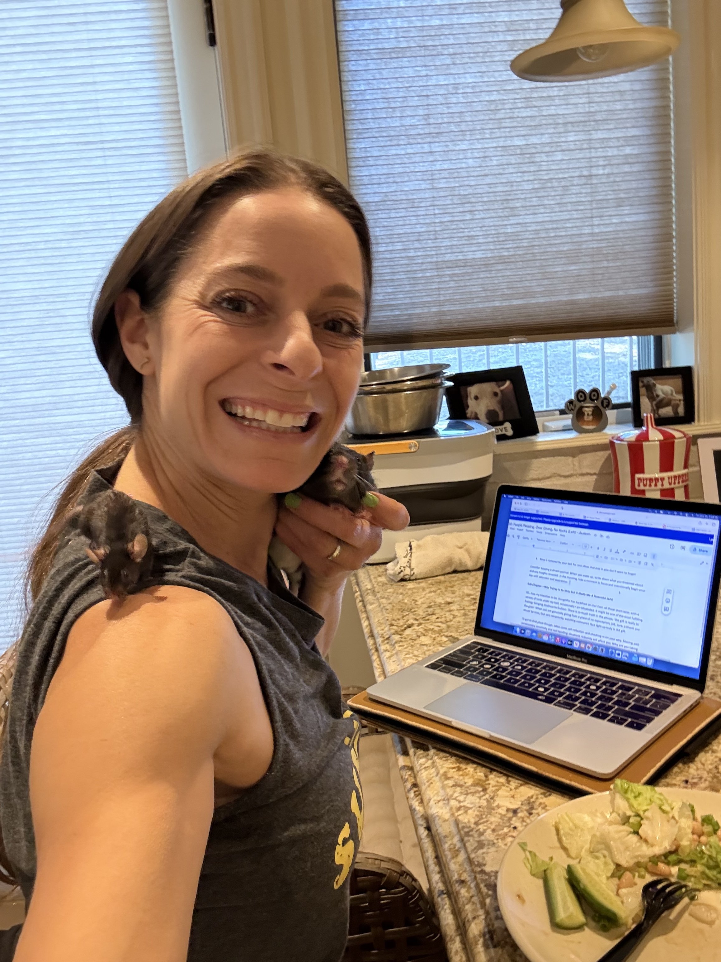 A woman with brown hair smiling while holding a small black puppy near her face. She is sitting at a kitchen counter with a laptop, a plate of food, and framed pictures of dogs in the background.