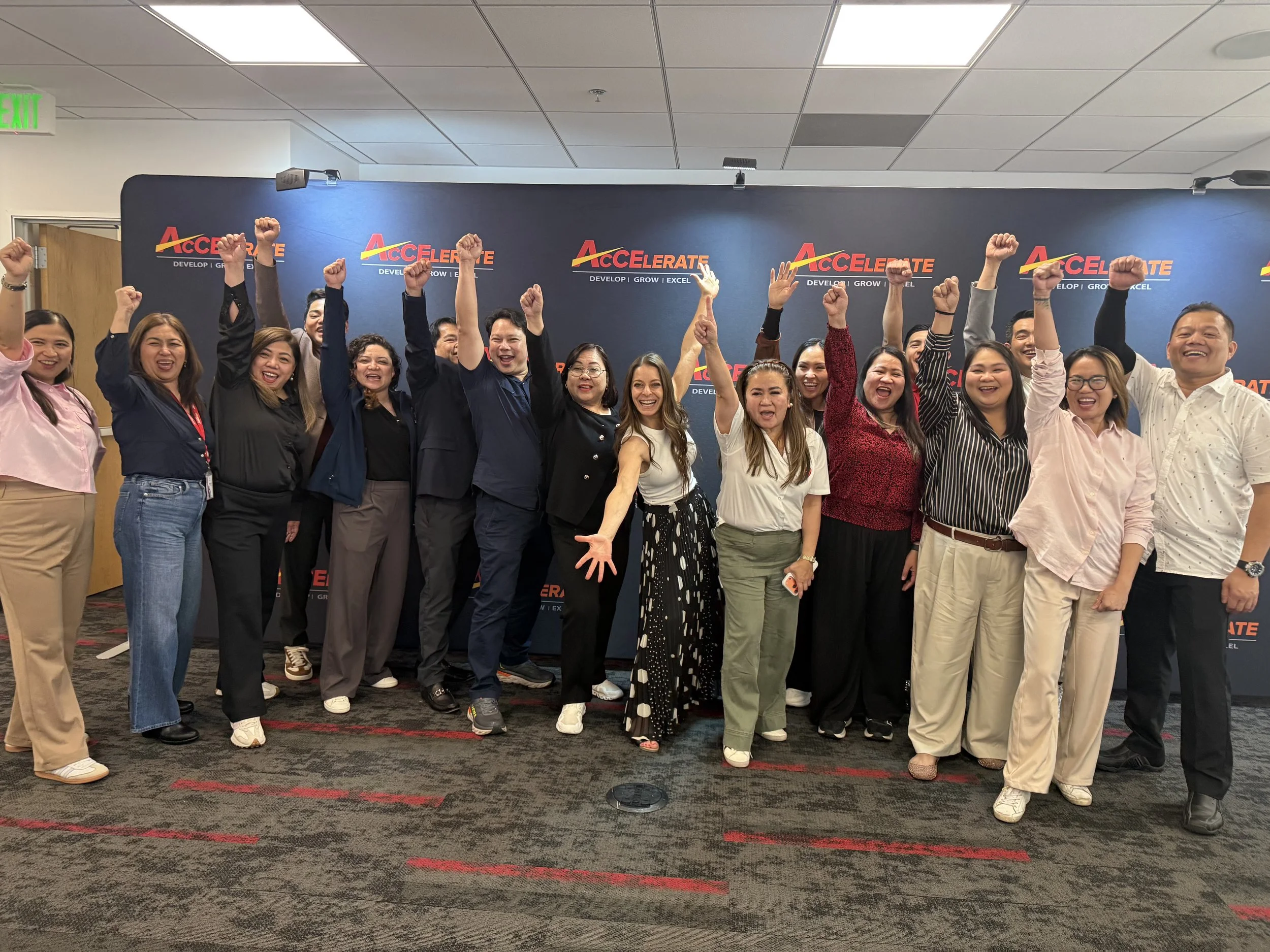 Group of people celebrating or cheering in front of a blue backdrop with the word 'ACCELERATE' and the slogan 'Develop, Grow, Excel'.