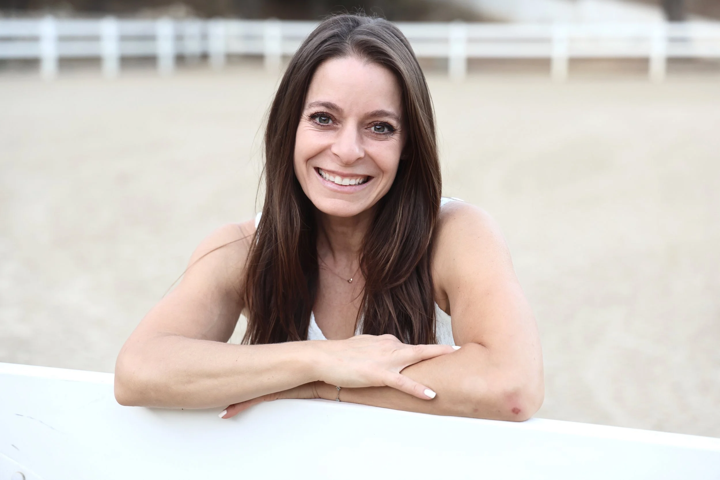 A woman with long brown hair and a bright smile sitting outdoors with arms crossed on a white surface, blurred background with a fence.