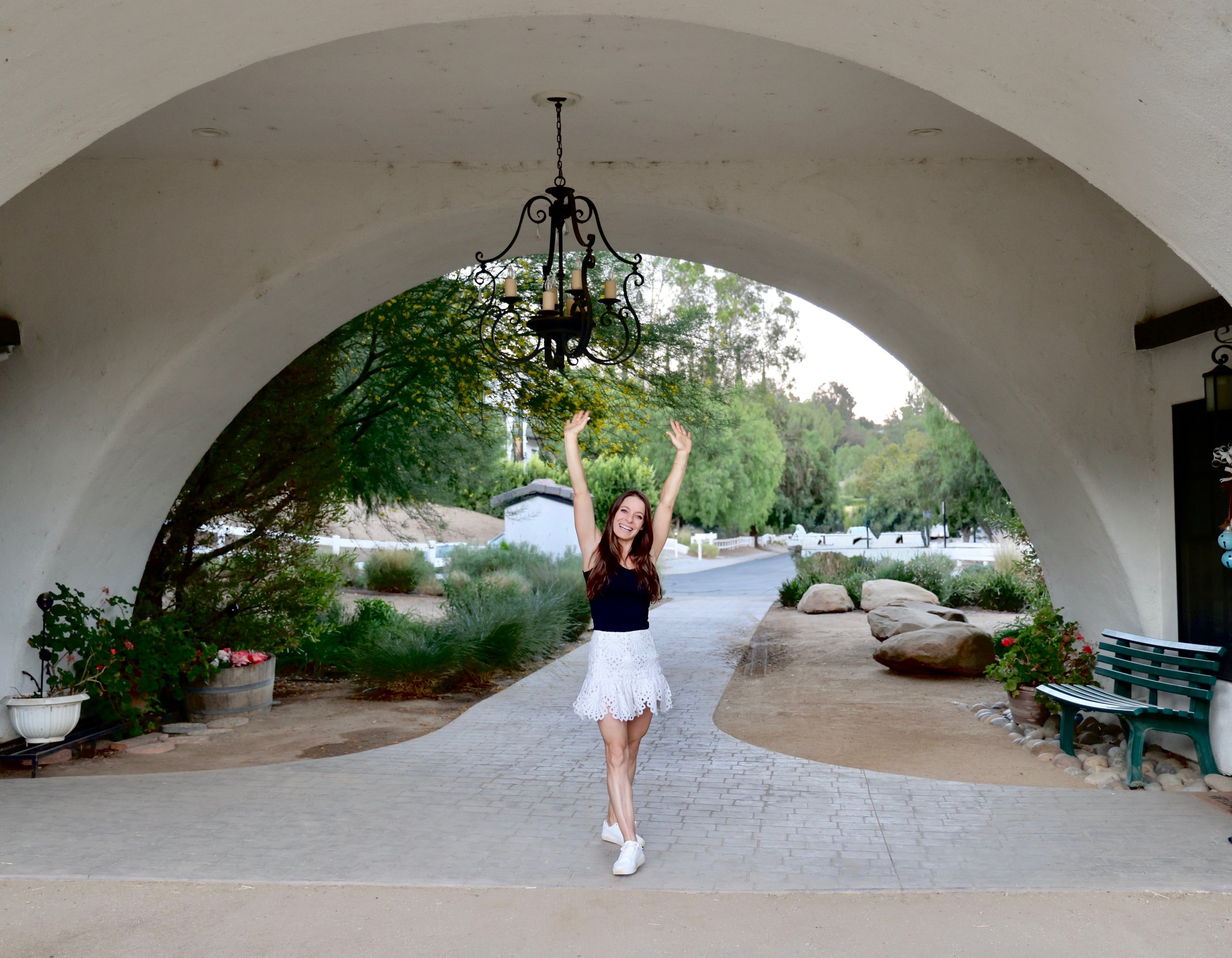 A woman in a white dress and white shoes standing under a large archway with her arms raised and smiling. There are trees and a pathway outside.