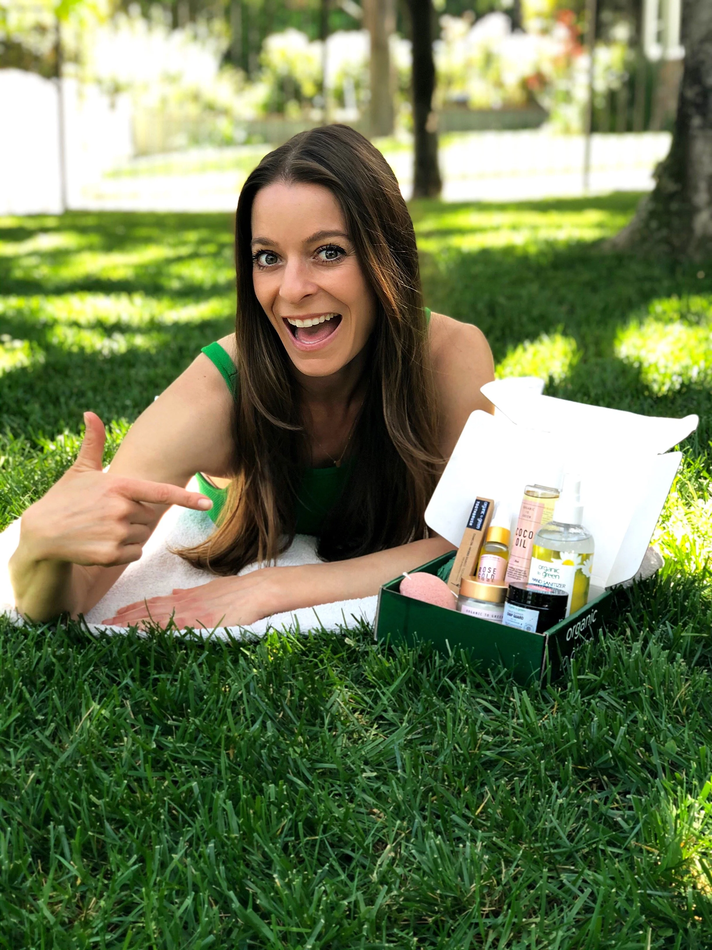 A woman with long dark hair lying on the grass in a park, smiling and pointing at a box of organic skincare products, with trees and a white fence in the background.
