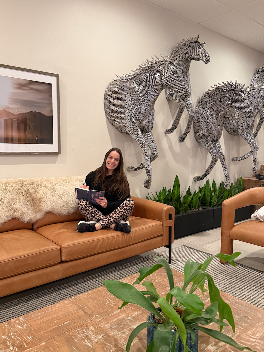 A woman sitting cross-legged on a tan leather sofa, smiling and holding an open notebook, in a room with decorative metal horse wall art, a large framed photograph, green potted plants, and wooden furniture.