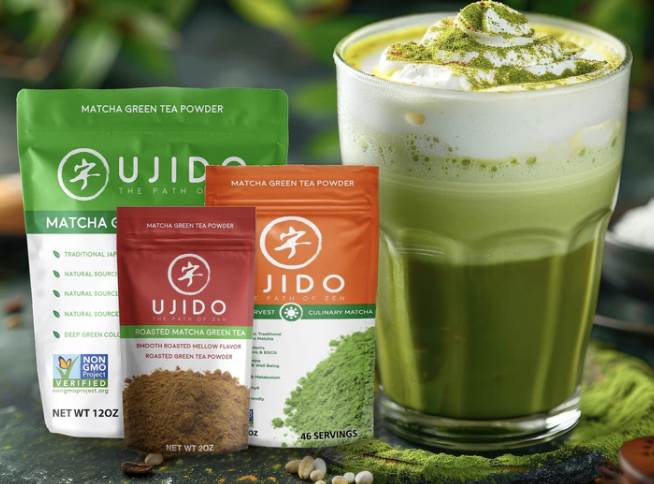 Three packets of Udido matcha green tea powder in green, orange, and red packaging beside a glass of prepared matcha latte topped with whipped cream and matcha powder, on a dark surface with a blurred green background.