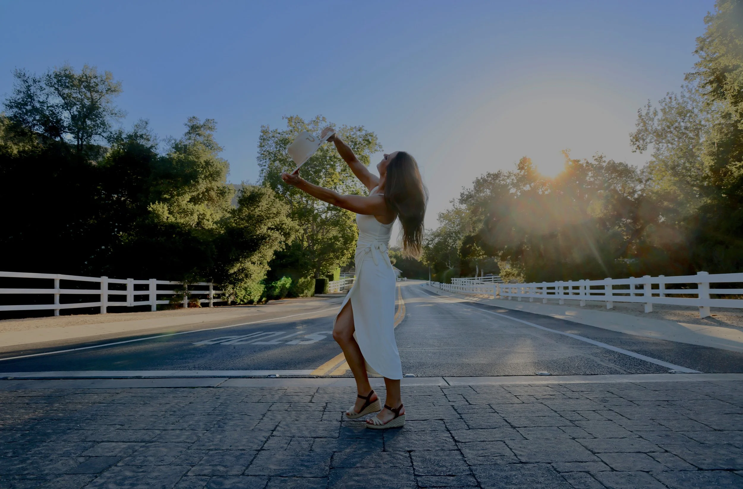 Woman in a white dress and wedge sandals dancing on a street with trees and a white fence, holding a watering can, with sunlight shining through the trees.