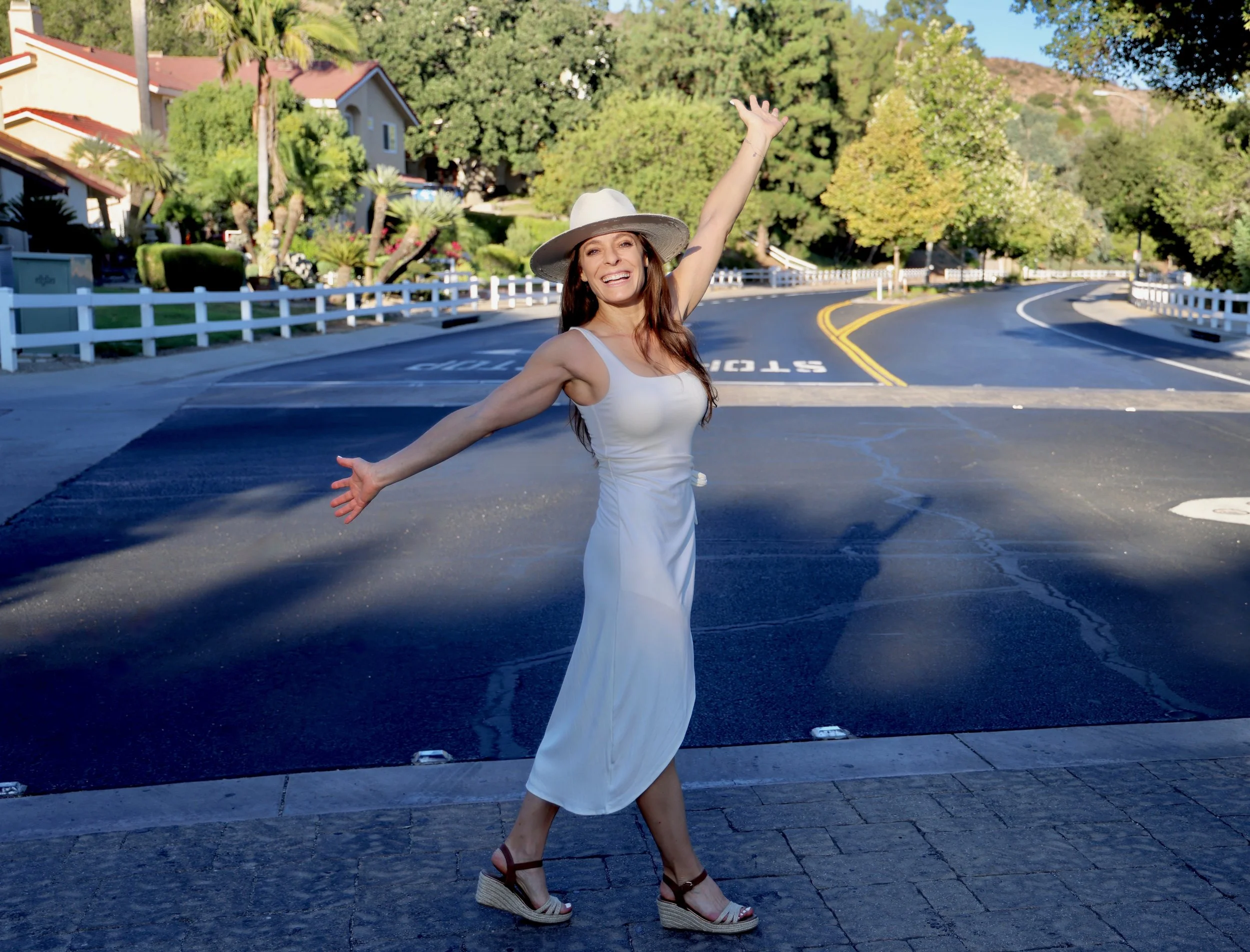 Woman in a white dress and large sunhat dancing or striking a joyful pose on a sidewalk in a residential neighborhood with green trees and houses in the background.