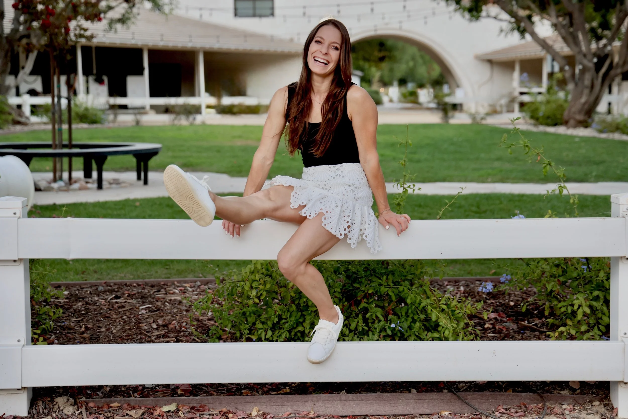 A woman sitting on a white fence smiling, wearing a black top, white eyelet skirt, and white sneakers in a park with trees and a bridge in the background.