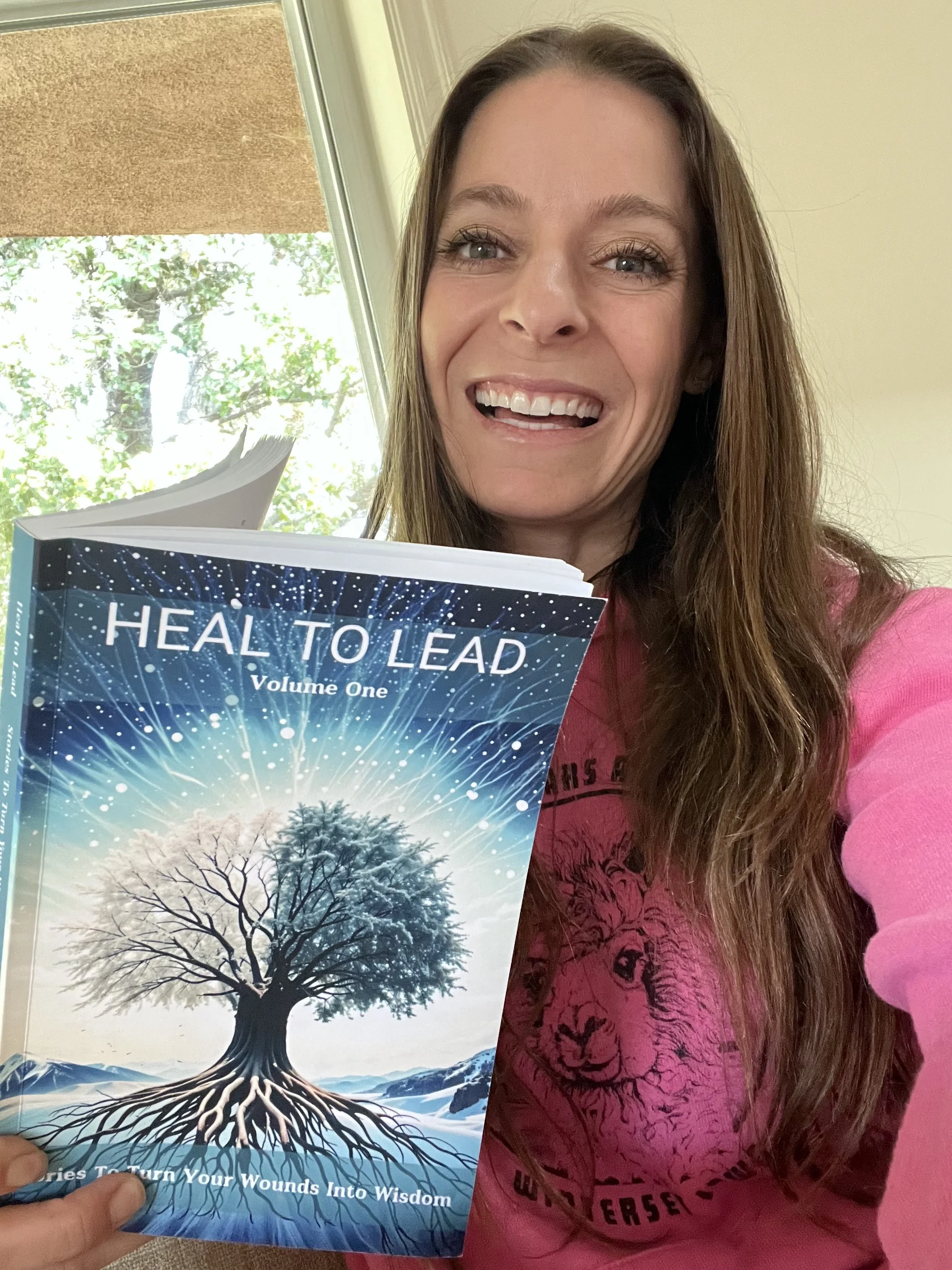 A woman with long brown hair and light skin, smiling while holding a book titled "HEAL TO LEAD: Turn Your Wounds Into Wisdom" in front of a window with green trees outside.