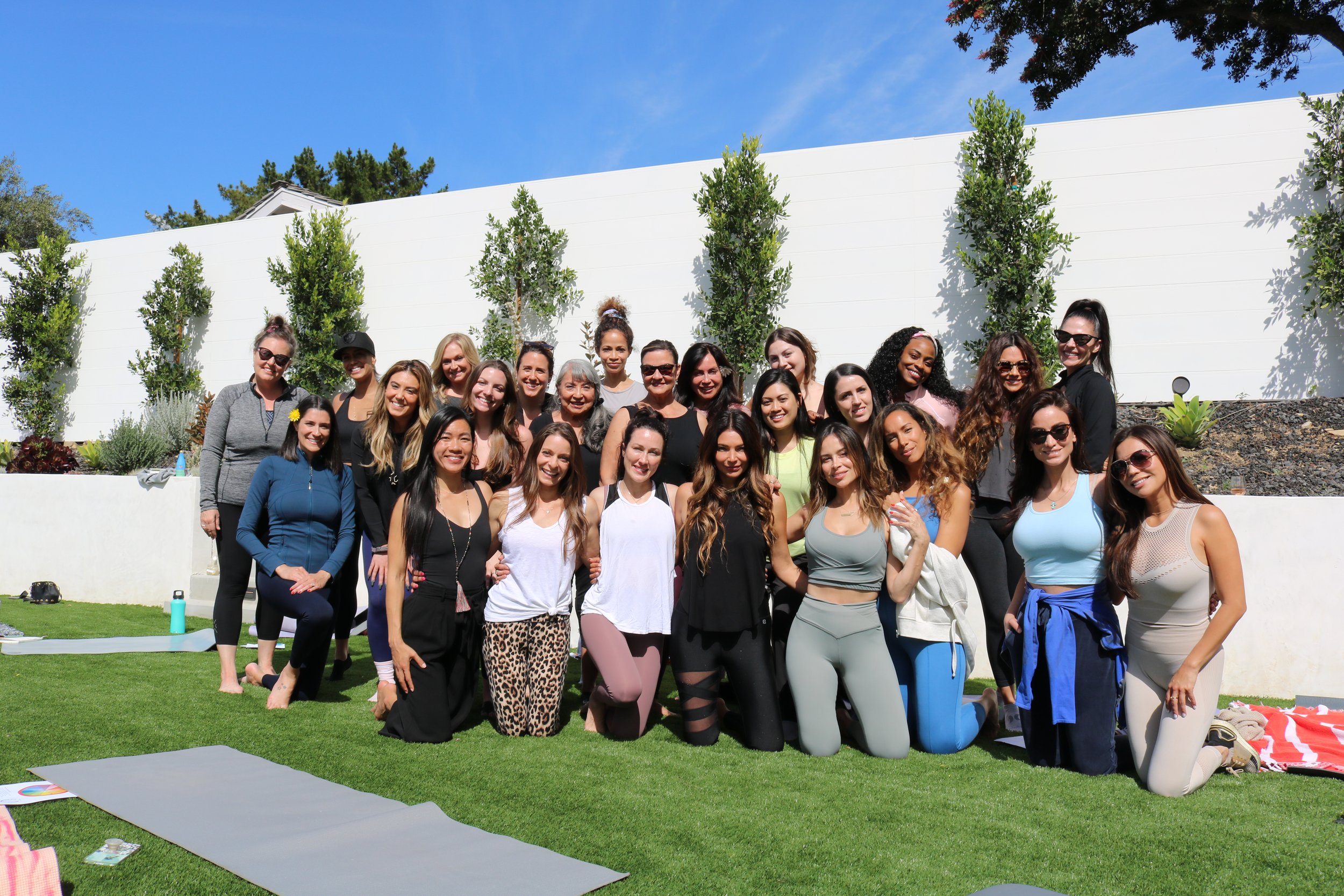 A large group of women of diverse backgrounds and ages posing outdoors on a sunny day in front of a white wall and green trees, smiling and dressed in casual athletic clothing.