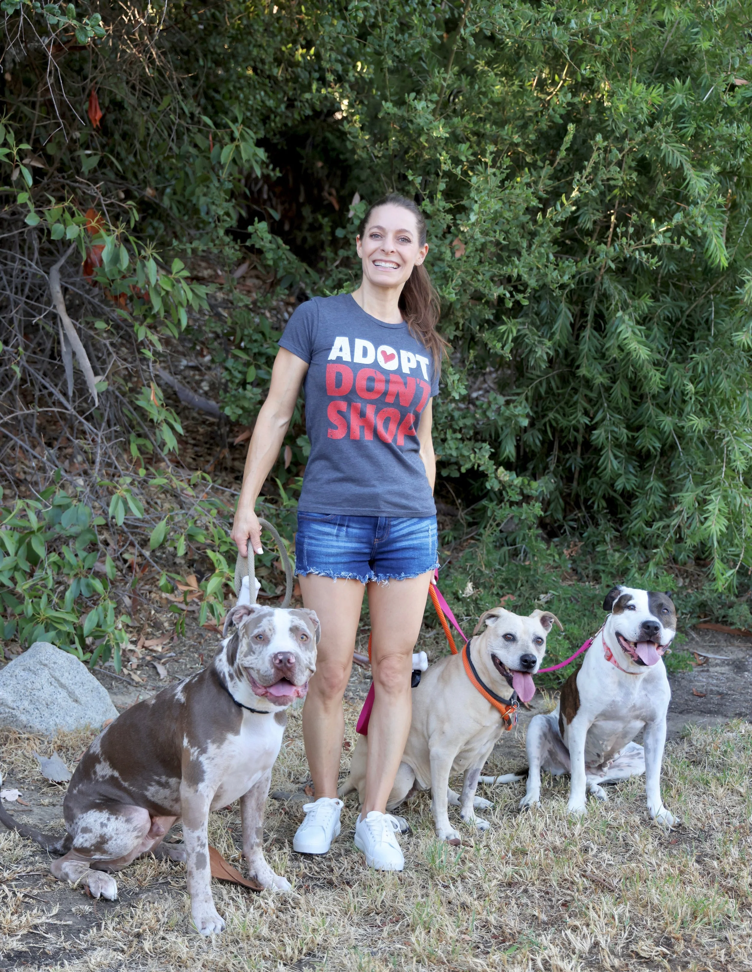 A woman smiling outdoors with four dogs, standing on grass and surrounded by green bushes.