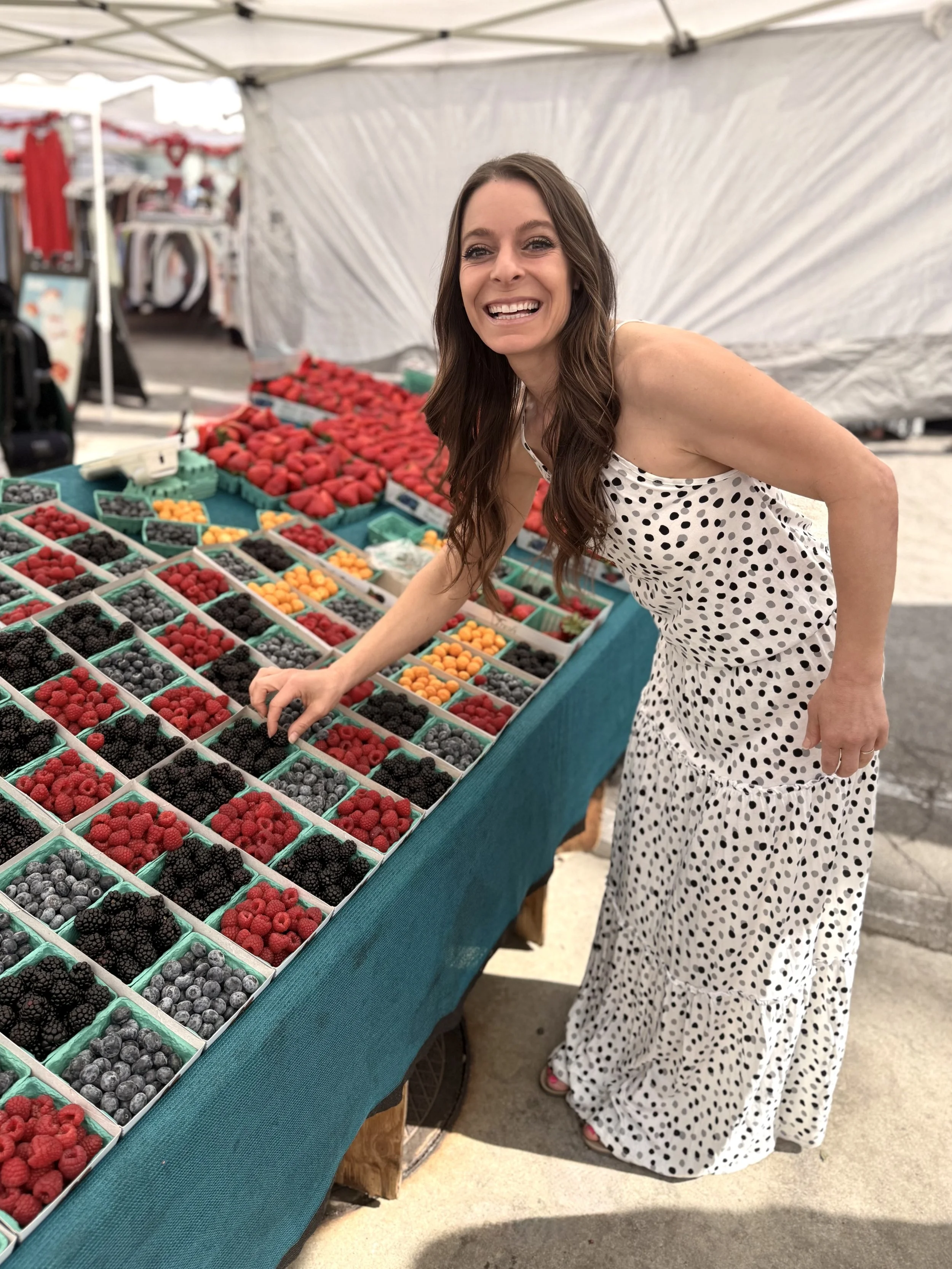 A woman in a polka dot dress is smiling next to a display of various fresh berries at an outdoor market.
