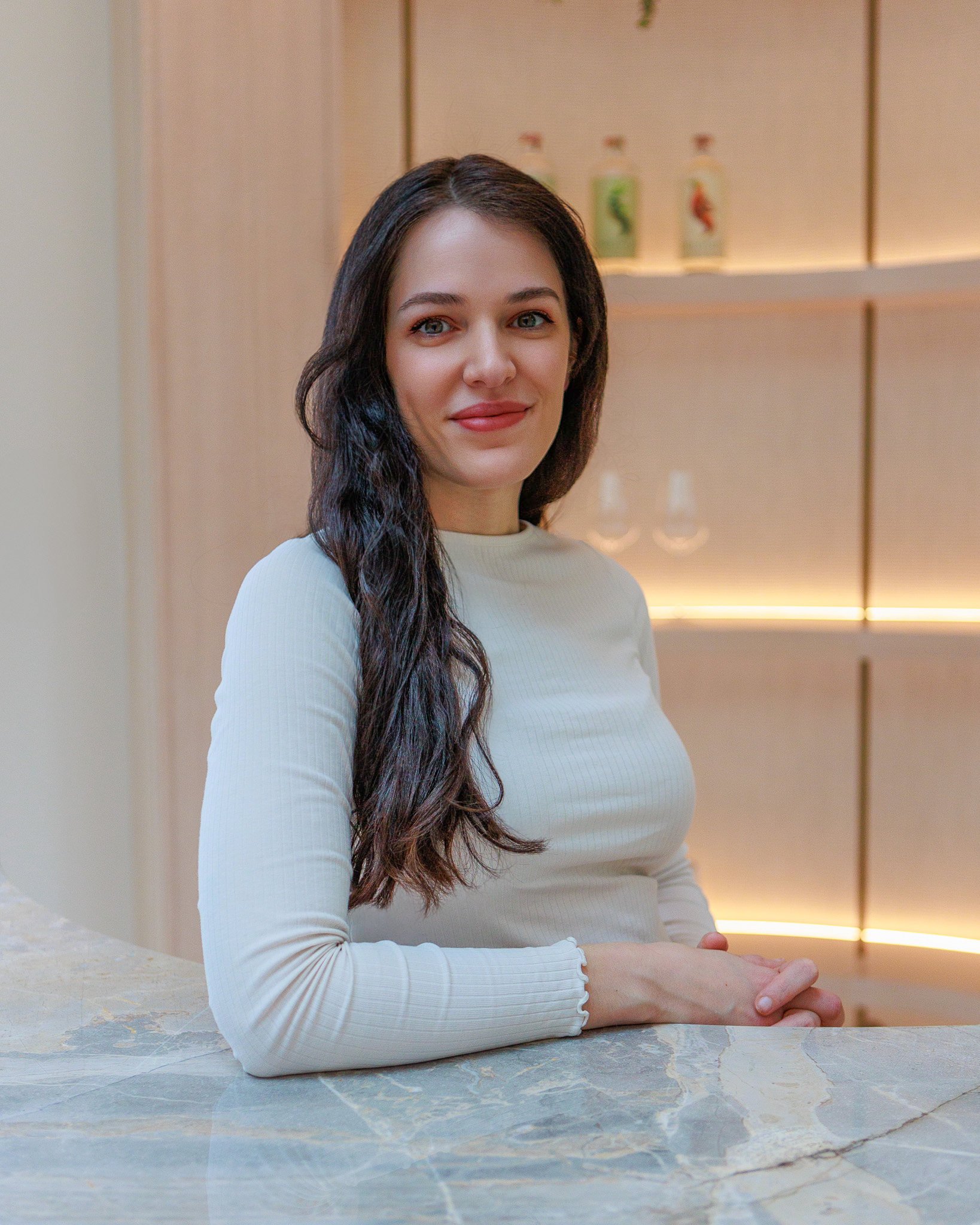 Person sitting at a table with a white top and long hair, smiling with arms crossed in a well-lit interior space.