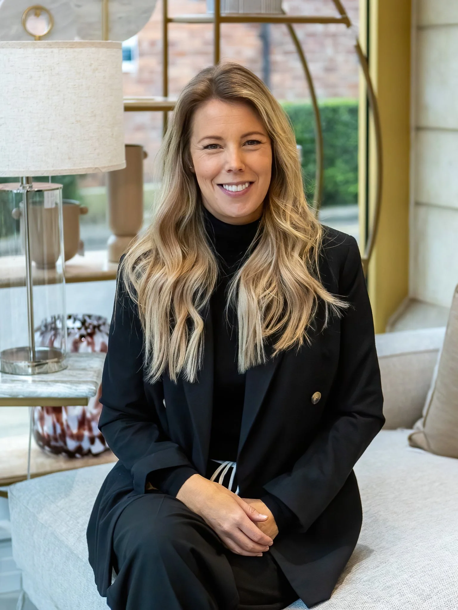 Person sitting indoors on a couch, wearing a black suit, with long blonde hair, smiling, next to a lamp.