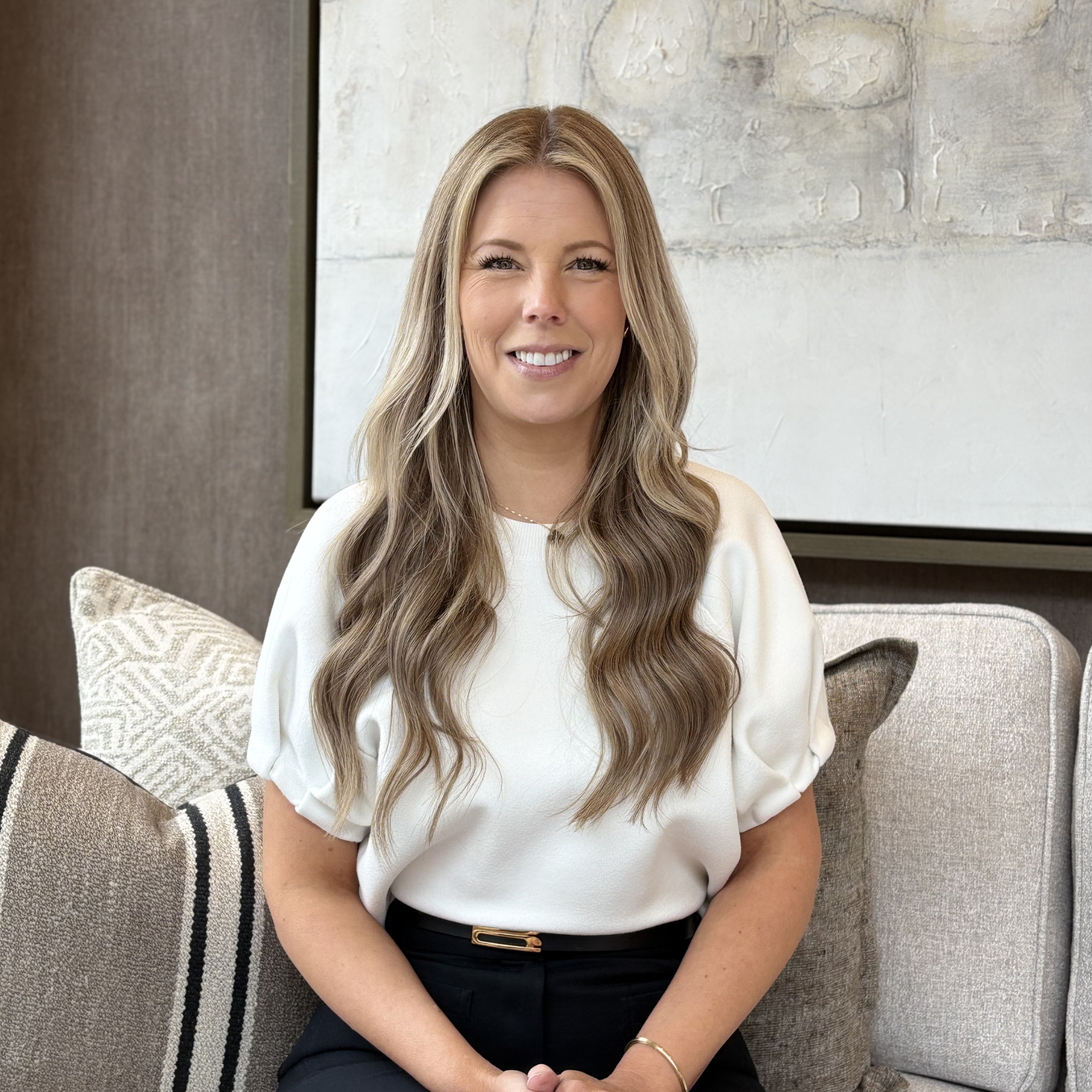 Person sitting indoors on a couch, wearing a black suit, with long blonde hair, smiling, next to a lamp.