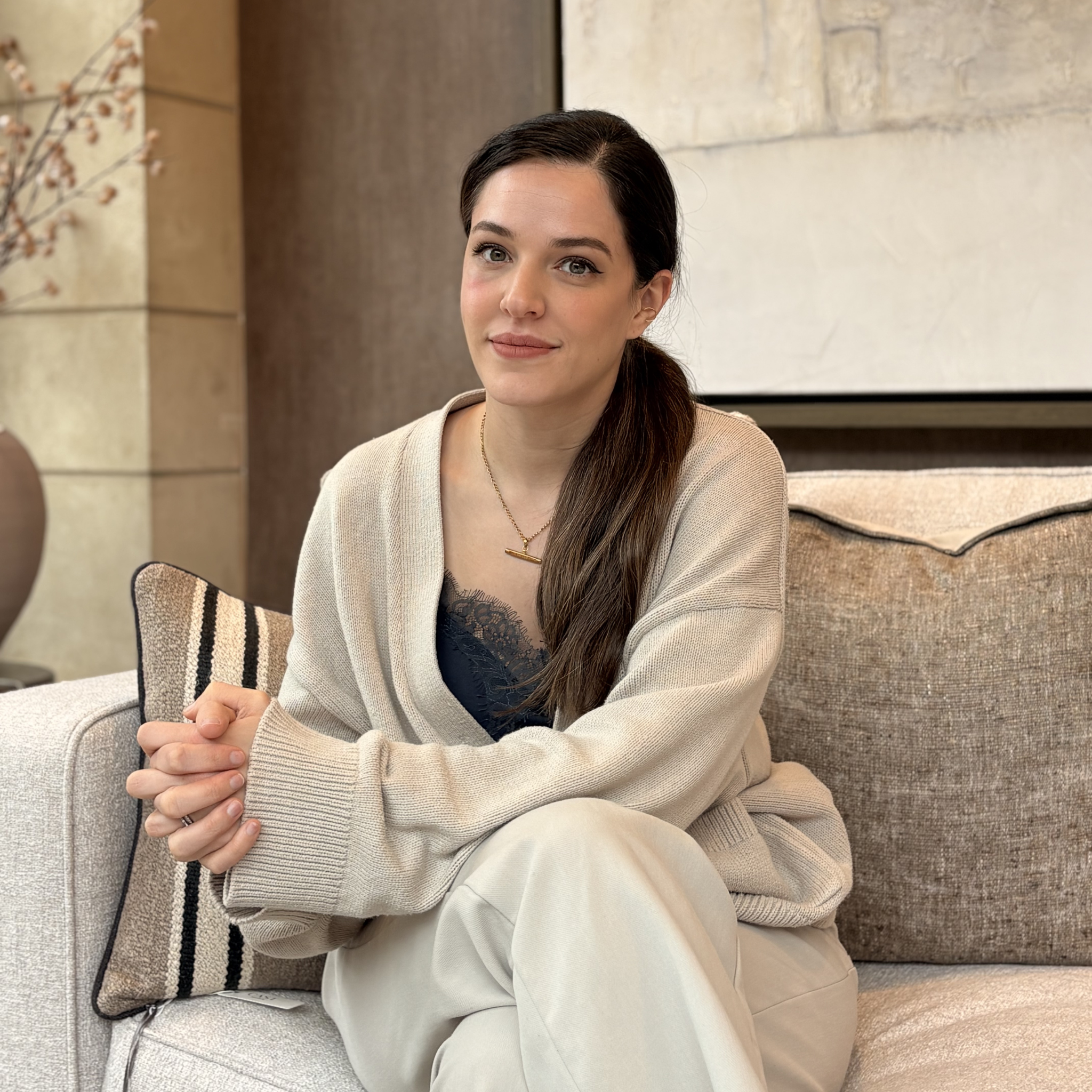Person sitting at a table with a white top and long hair, smiling with arms crossed in a well-lit interior space.