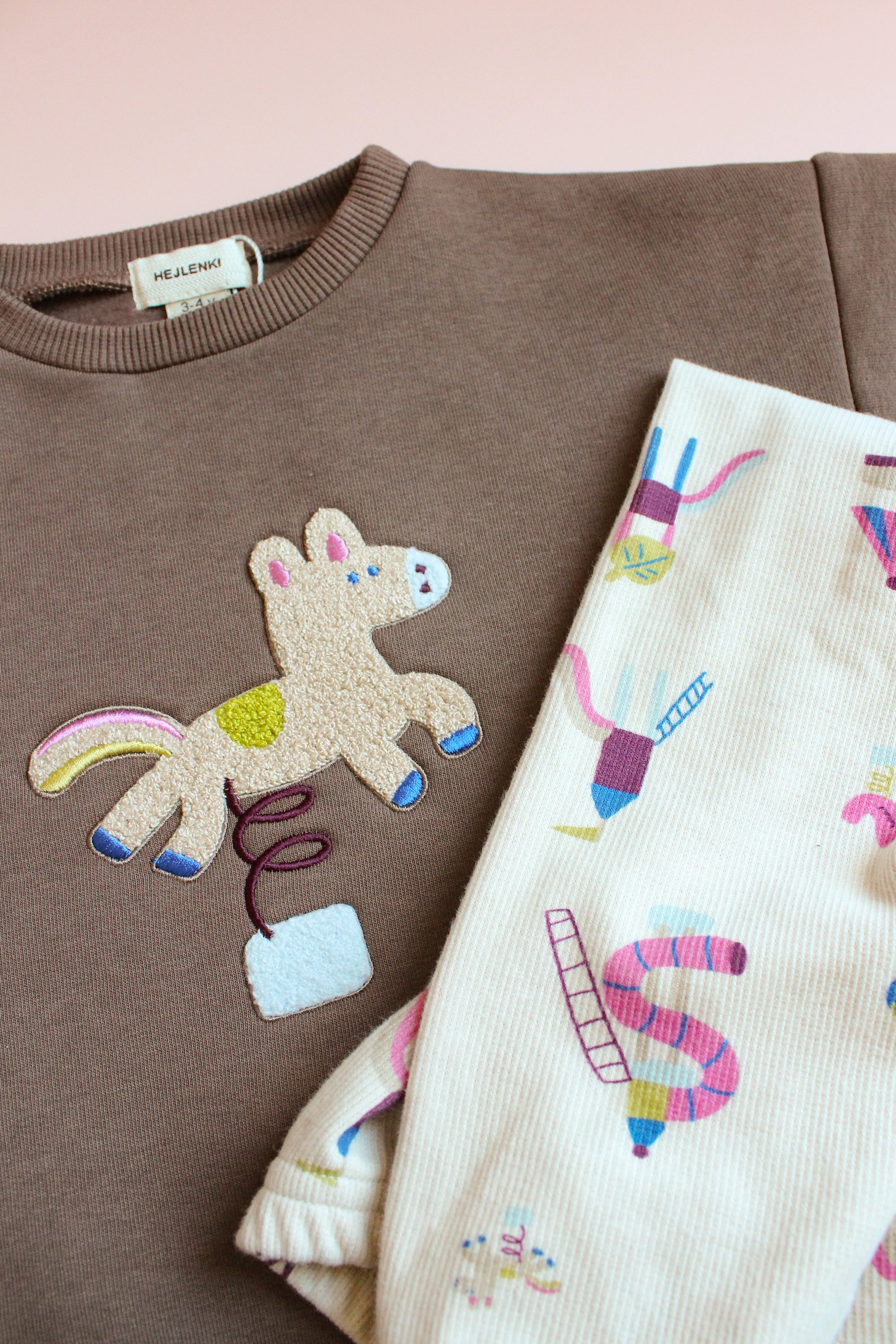 Deux vêtements pour enfants, un en brun avec une broderie d'une licorne dans un pot de fleurs et un en blanc avec un motif coloré de sucres d'orge, bâtons de bonbon, et autres friandises.