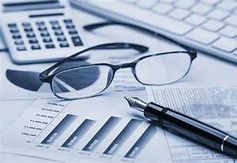 Glasses, a pen, and financial documents with bar charts on a desk, with a calculator and keyboard in the background.