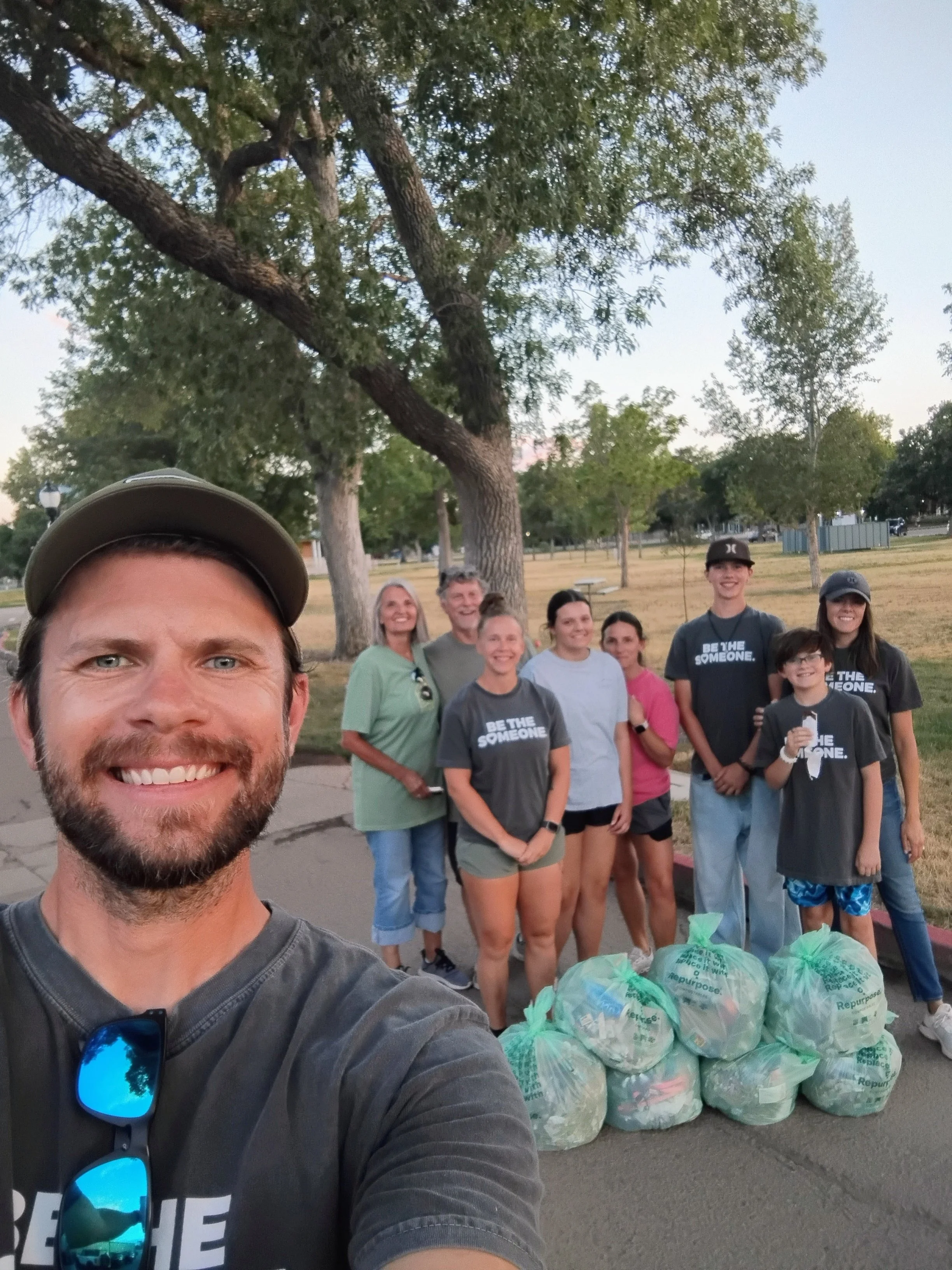 Three people, a man and two boys, participating in a park cleanup. The man is taking a selfie, wearing sunglasses and a gray t-shirt that says 'BE THE SOMEONE.' The boys, also in gray T-shirts with the same message, are holding trash pickers, standing near white buckets and black garbage bags on a dirt path in a dry, grassy area.