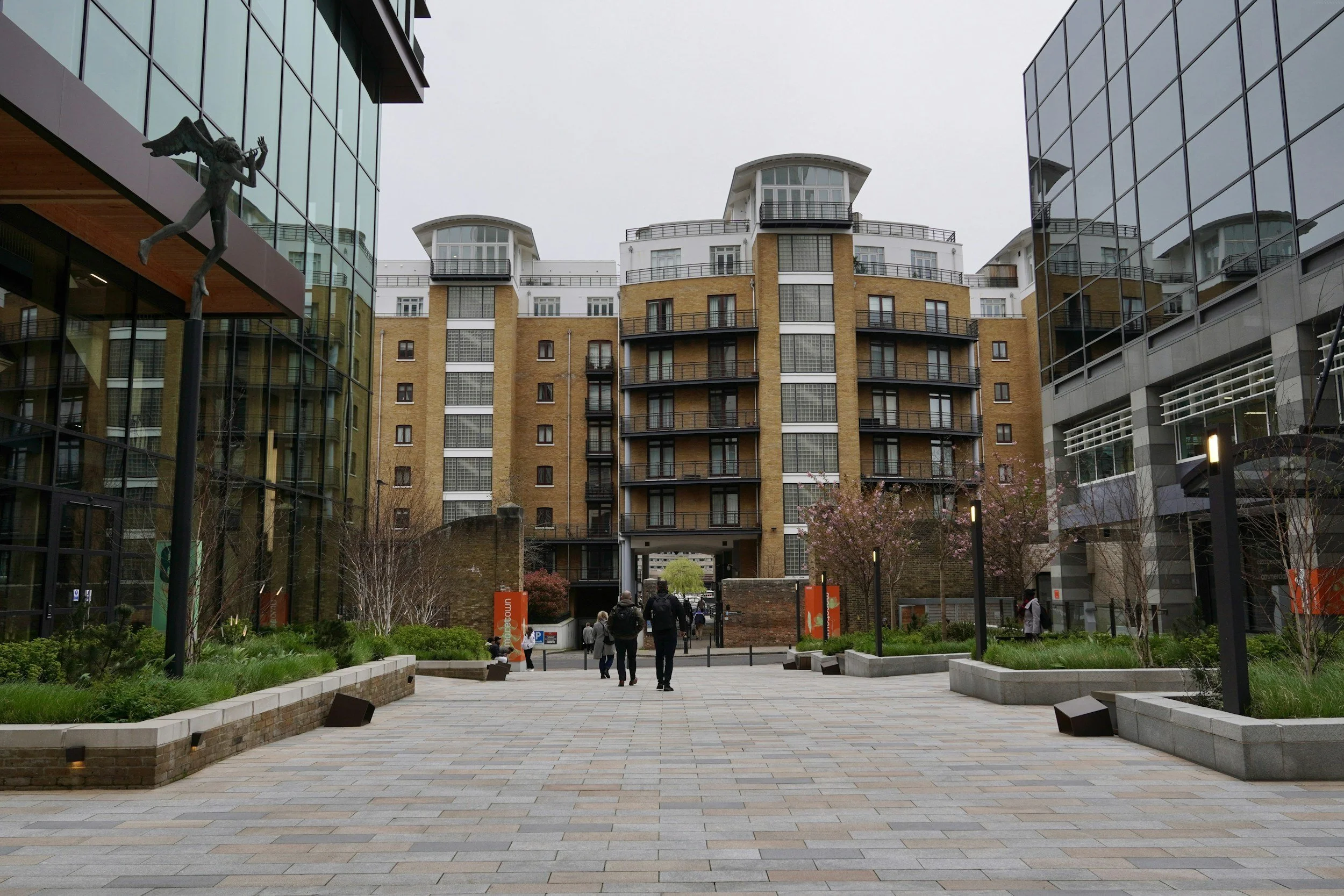 People walking through a modern urban courtyard with landscaped planters, surrounded by high-rise apartment buildings with glass facades and brick exteriors