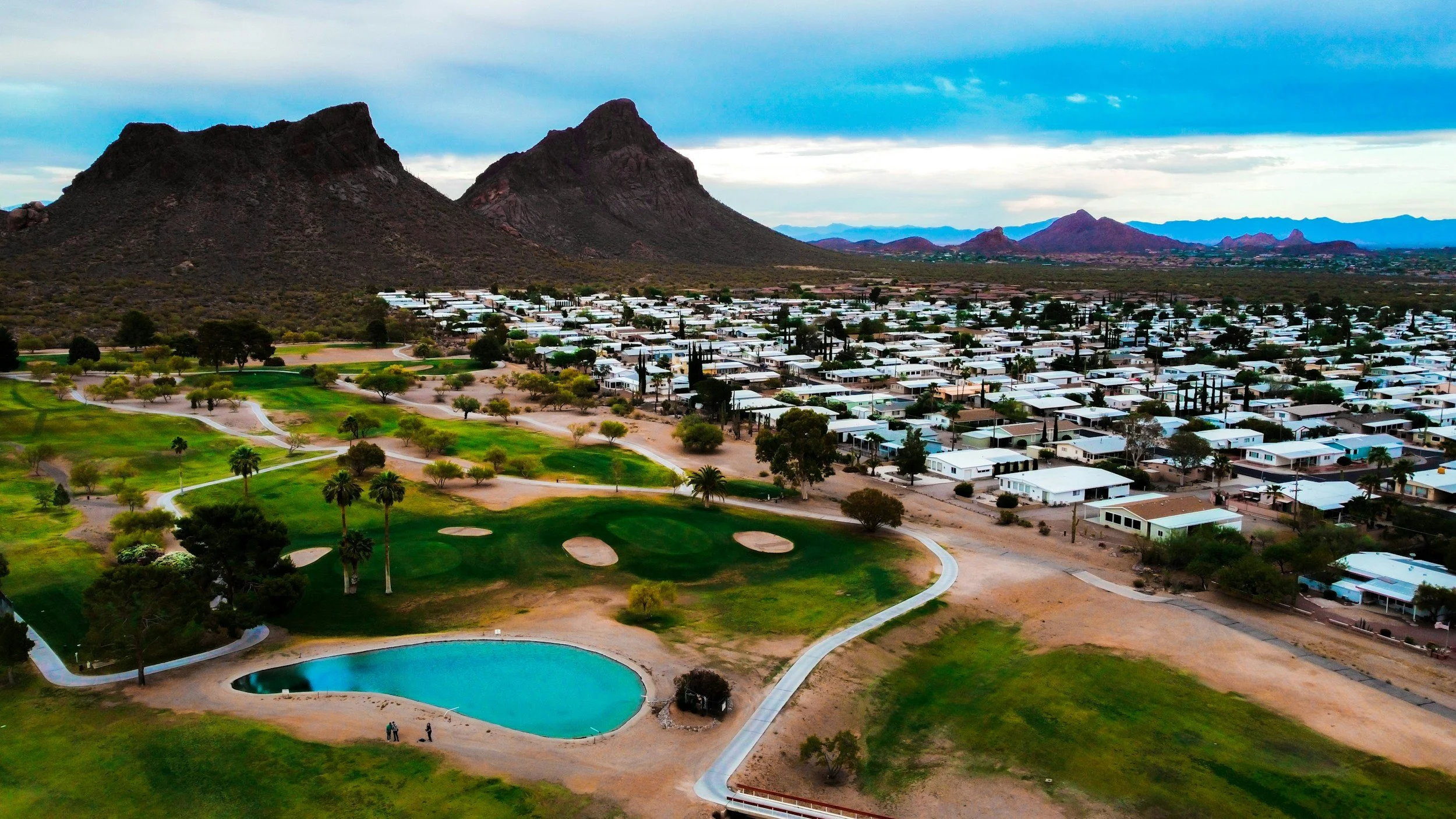 Aerial view of a golf course with a large swimming pool, surrounded by trees, with a residential neighborhood and mountains in the background.