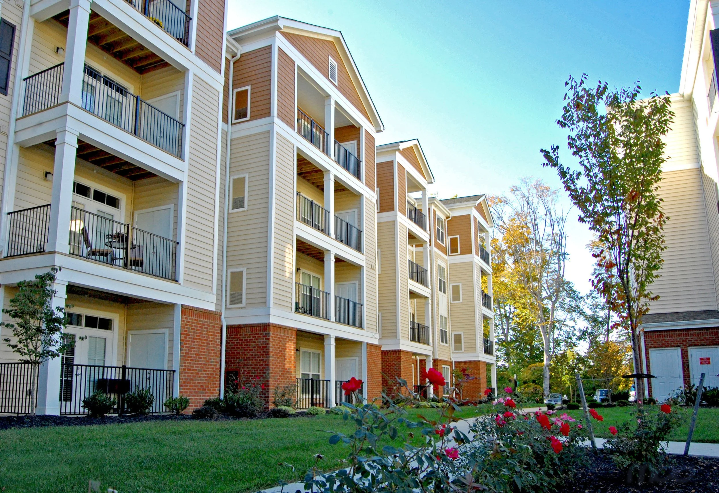 Multi-story residential apartment building with balconies, brick accents, and landscaped yard with flowers and trees.
