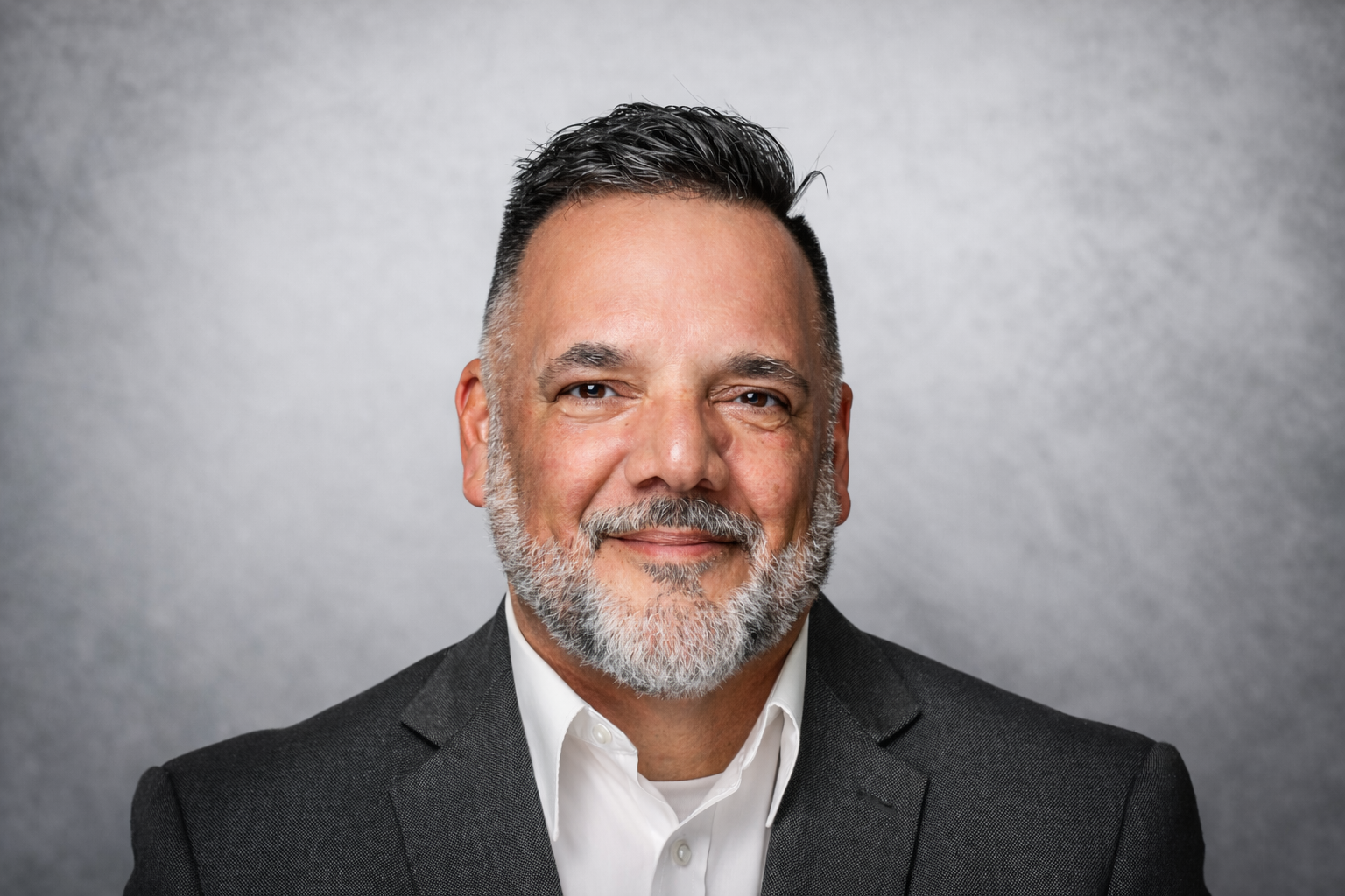 A headshot of a middle-aged man with gray beard and hair, wearing a dark gray suit and white shirt, smiling against a gray textured background.