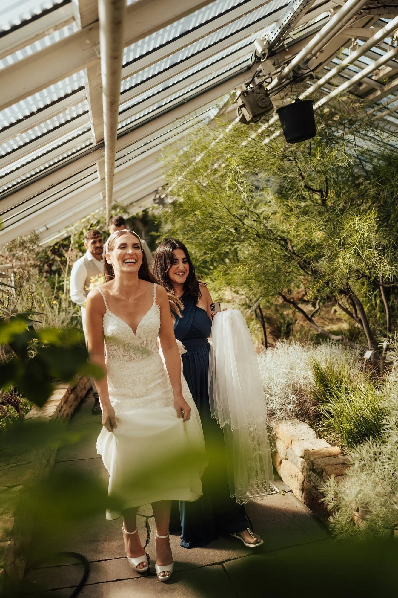 Bride smiling in a glasshouse full of plants, with two guests walking behind her