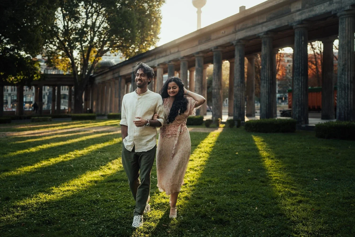 Couple walking hand in hand in low sun by a colonnade, the TV tower behind