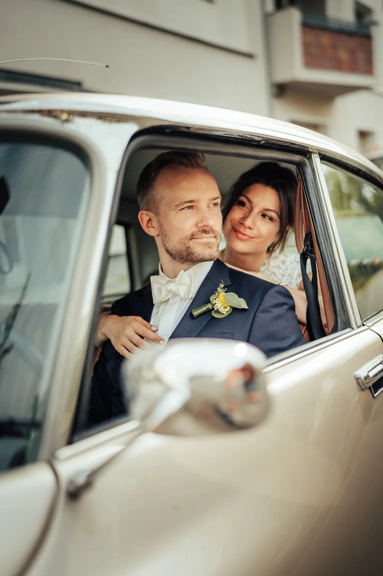 Bride and groom smiling from the window of a vintage car on their wedding day