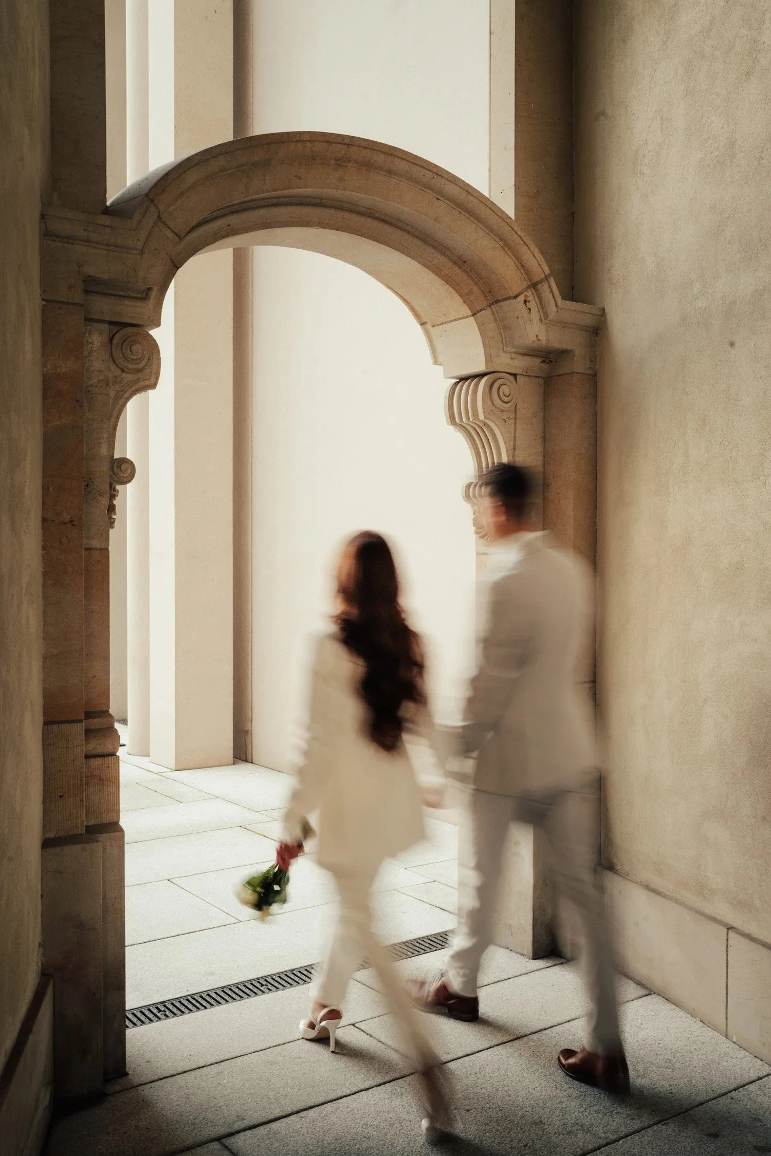 Couple in pale suits walking through a stone archway after their wedding
